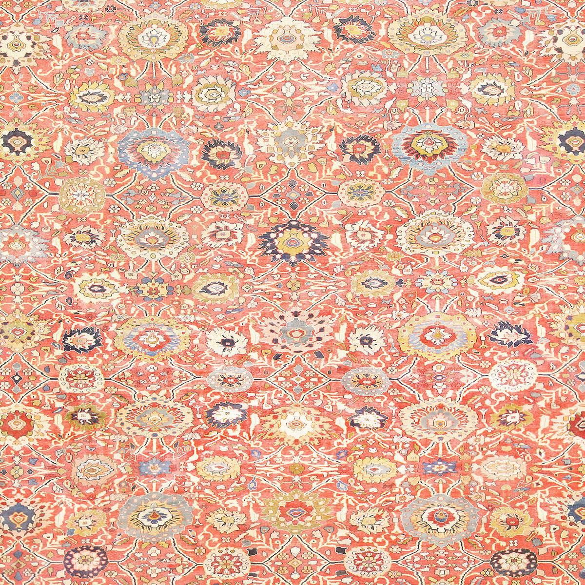A lively pattern fills this antique Persian rug, covering the large oversized red-orange backdrop with an even array of blossoms in yellow, white, red and blue. Dark blue accents and lines give the light hues of this antique Persian Sultanabad rug