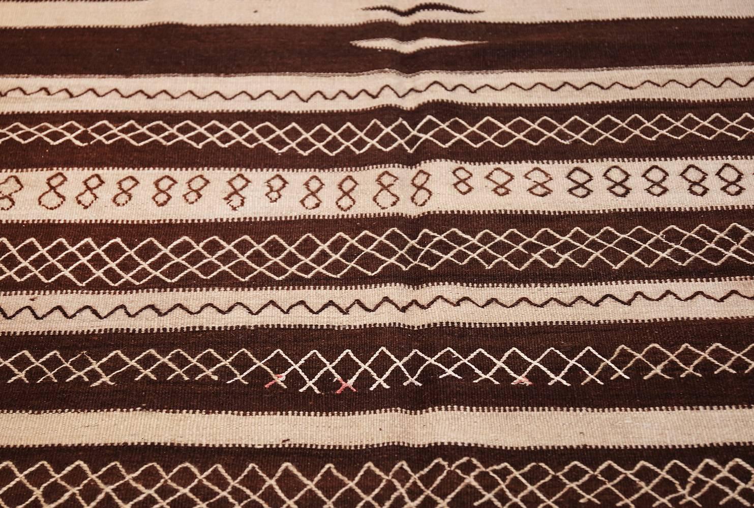 Drawn in a bold, graphic style, this vintage Moroccan Kilim depicts a strong pattern of walnut brown and ivory stripes that is embellished with tiny latticework fences, almond-shaped windows and comb motifs with extended fingers. This foundation of