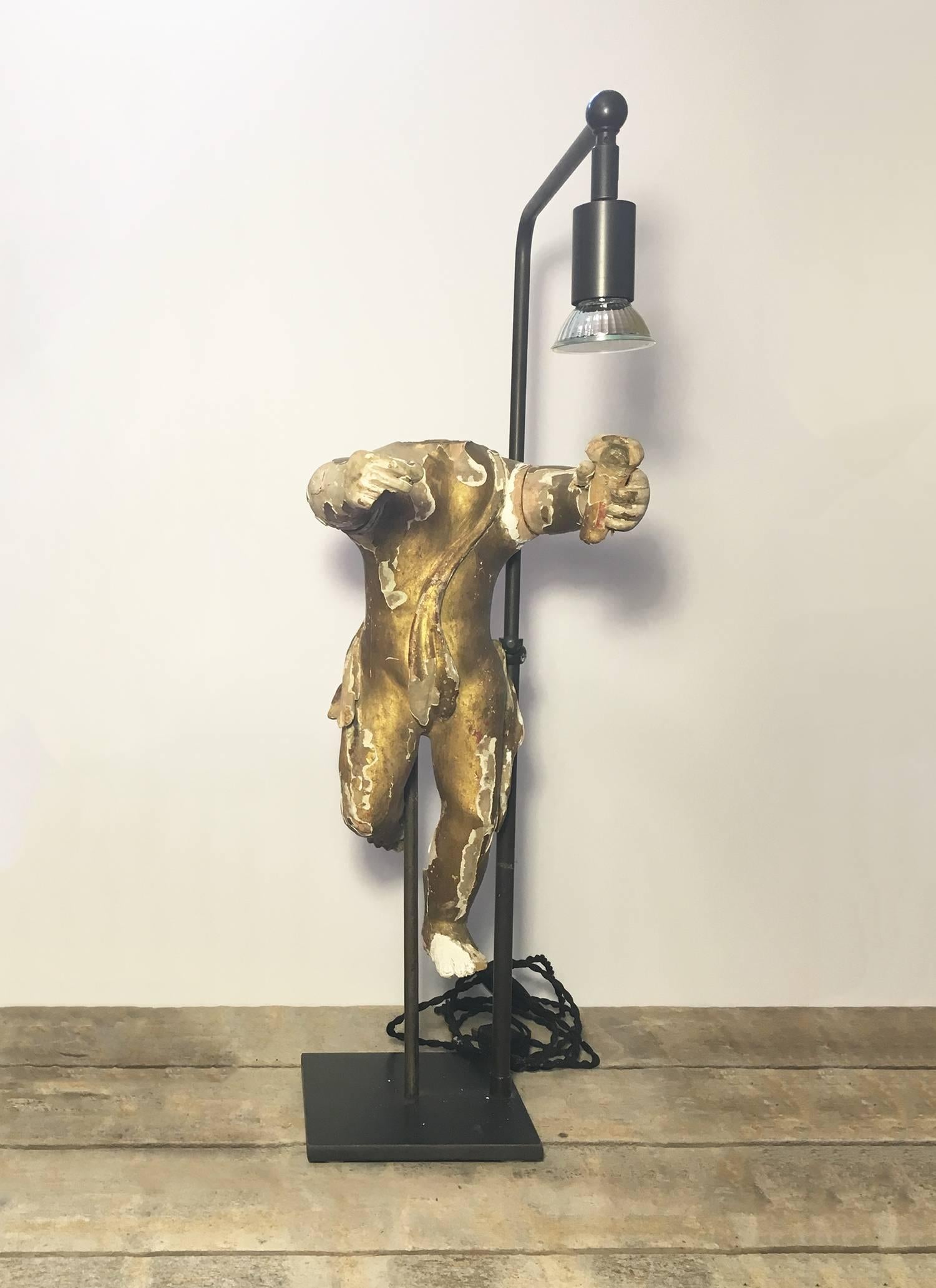 Wood, plaster and gilt figurine lamp depicting a putti or cherub holding a torch. 

Carved wood figure with layer of gilded plaster. Mounted on a black museum base with an adjustable light, wired for United States standard two prong outlet.