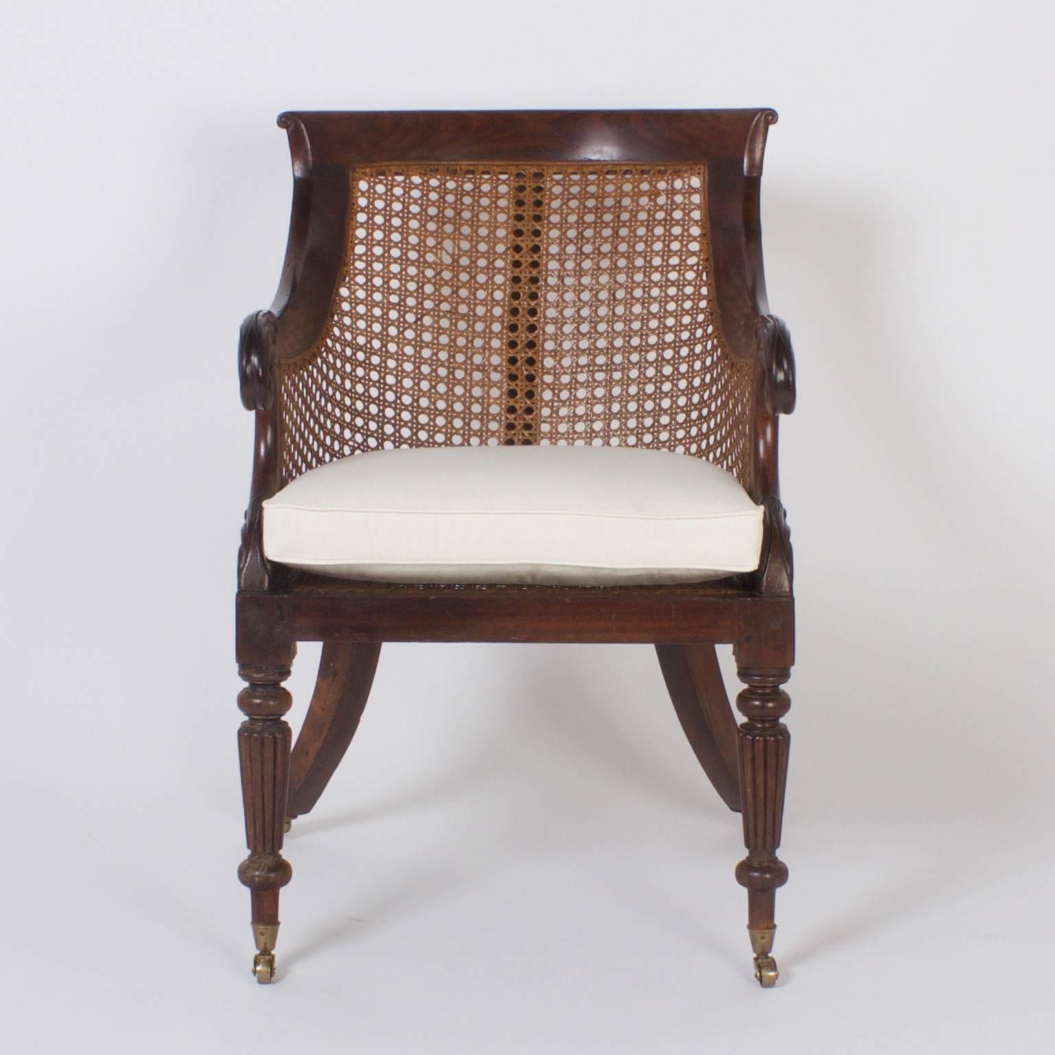 Rare pair of Regency style English library chairs crafted in mahogany and hand caned. These armchairs have a relaxed yet sophisticated design with carved acanthus arms, rounded backs, turned and reeded front legs and splayed back legs all set on