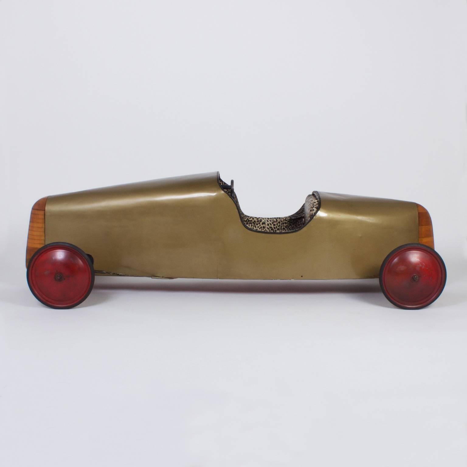 Here is a rare piece of Americana, a Mid-Century soap box derby racing car with sophisticated craftsmanship and classic form. The sleek body is gold lacquer. The interior is an unexpected leopard print vinyl. The front, rear and axel support