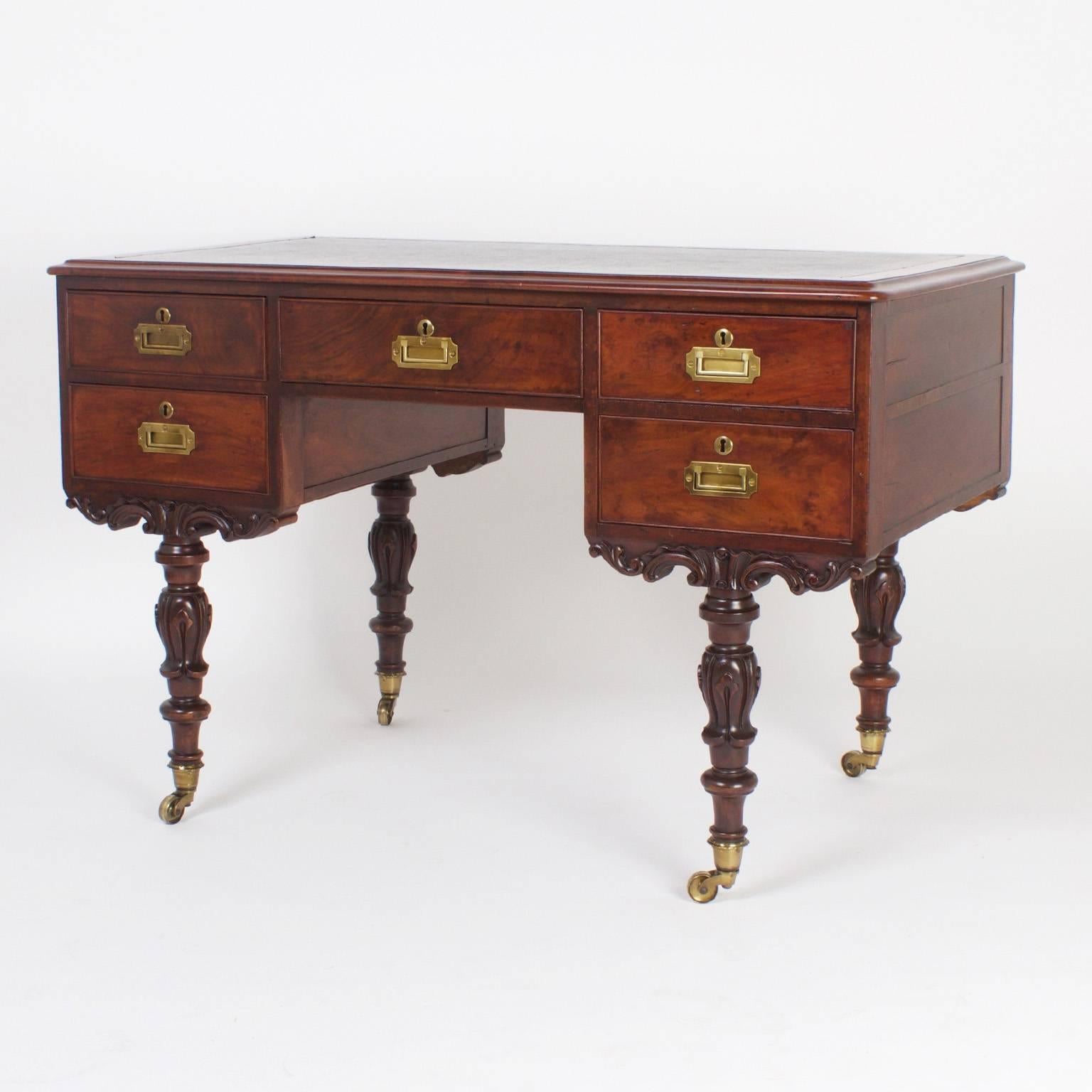 Rare and unusual Georgian style antique mahogany desk featuring a black leather top and inset campaign style brass hardware. The legs are off set from the corner and have a carved acanthus theme with lathe turned rings, all over brass casters.