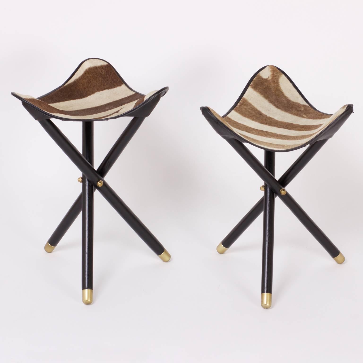 Dashing group of four of Mid-Century portable zebra stools or benches with folding tripod ebonized bases tipped with brass. One stool has a brown leather lining, note picture. Sold as a group for $5,200.00 or individually for $1,400.00 each.