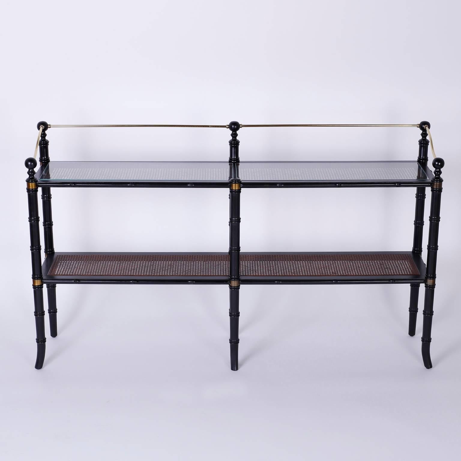 Campaign and Regency style two-tiered set of caned shelves or étagère with an elegant ebonized faux bamboo frame punctuated by brass cuffs and having a Classic brass gallery. The shelves are caned, glass protection on the upper tier.\
     