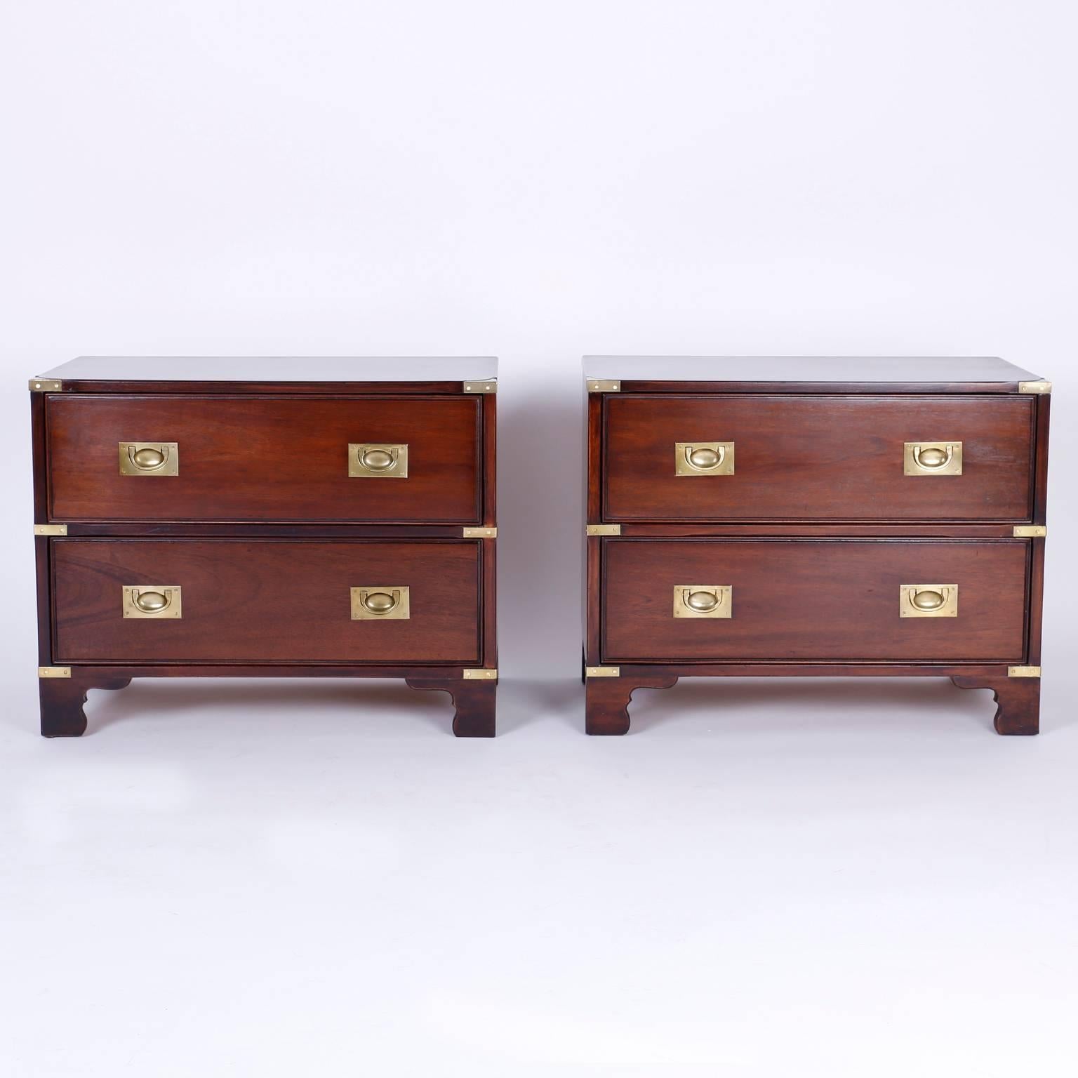 Dapper pair of Mid-Century mahogany nightstands or two-drawer chests with Campaign style and brass hardware set on bracket feet. 

