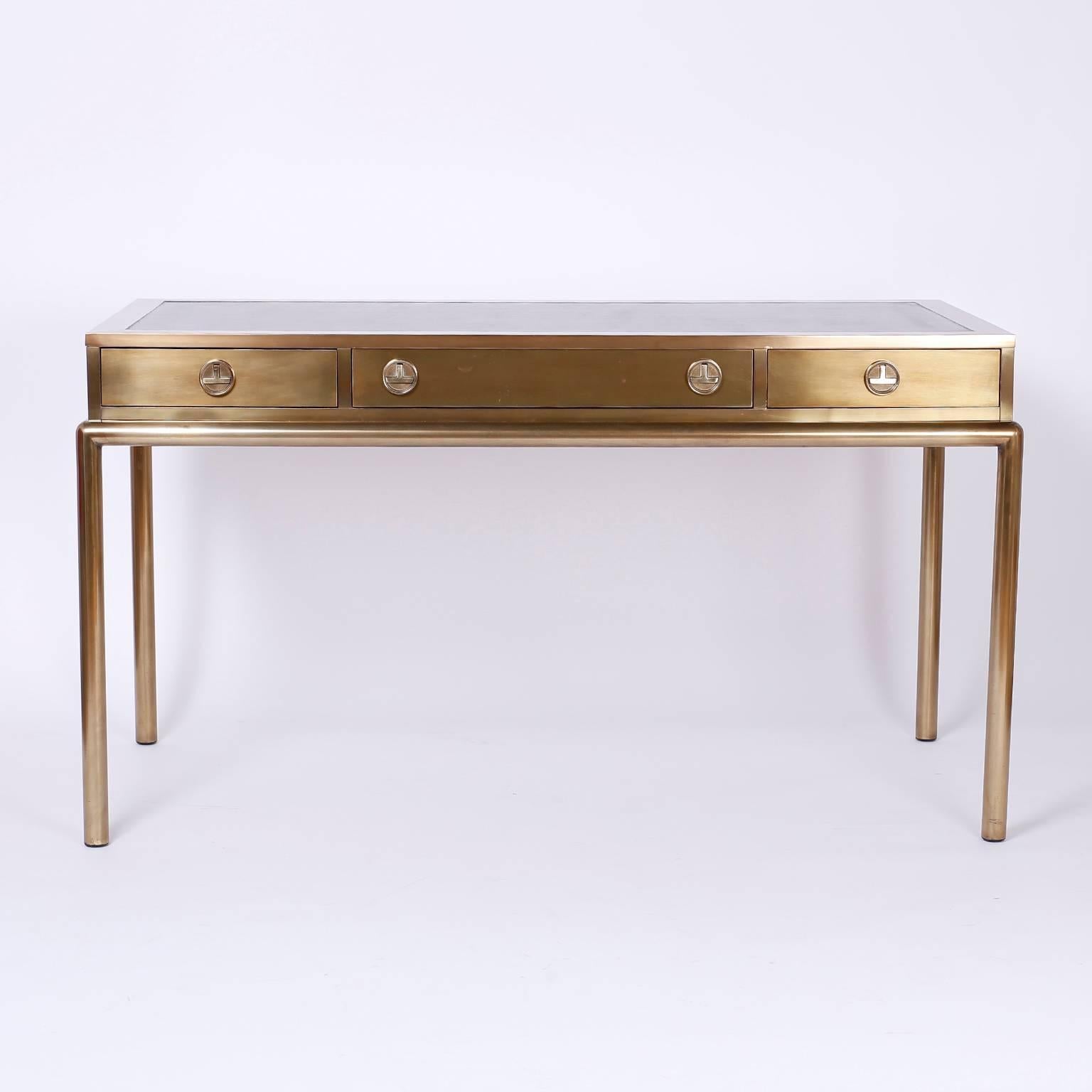 Chic brass bureauplat or desk with a clean no nonsense form, and a chinoiserie vibe. The top has a brown leather surface. The drawer pulls have stylized modern Campaign influences. The round legs complete the sleek design. The brass is lacquered for