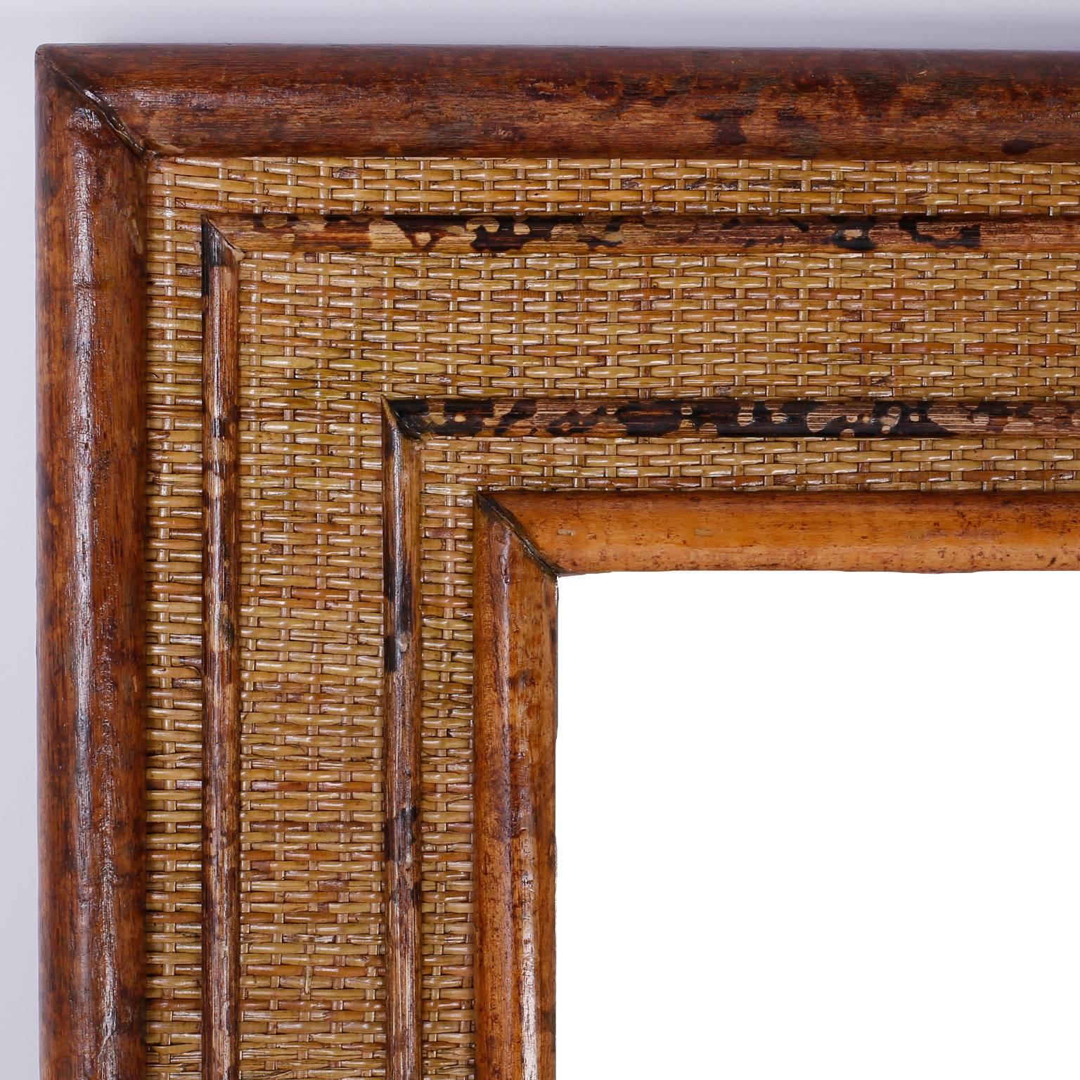 Dashing British colonial style beveled mirror with frame that is composed with faux finished bamboo and reed in geometric designs over a woven rattan background.
   