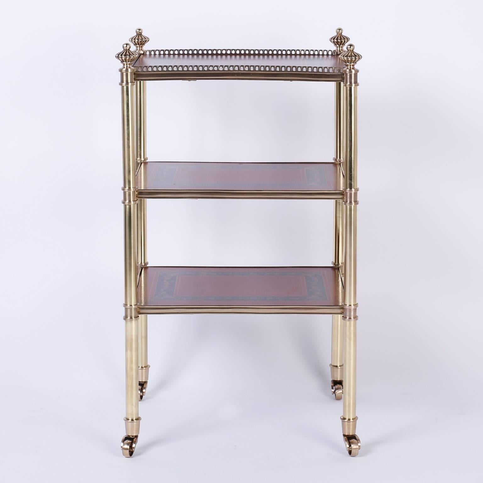 Three-tiered serving or display stand with a dashing polished and lacquered brass frame topped, stylized finials and rolling on brass casters. The three shelves are decorated with tooled and painted red leather, in a classic design.