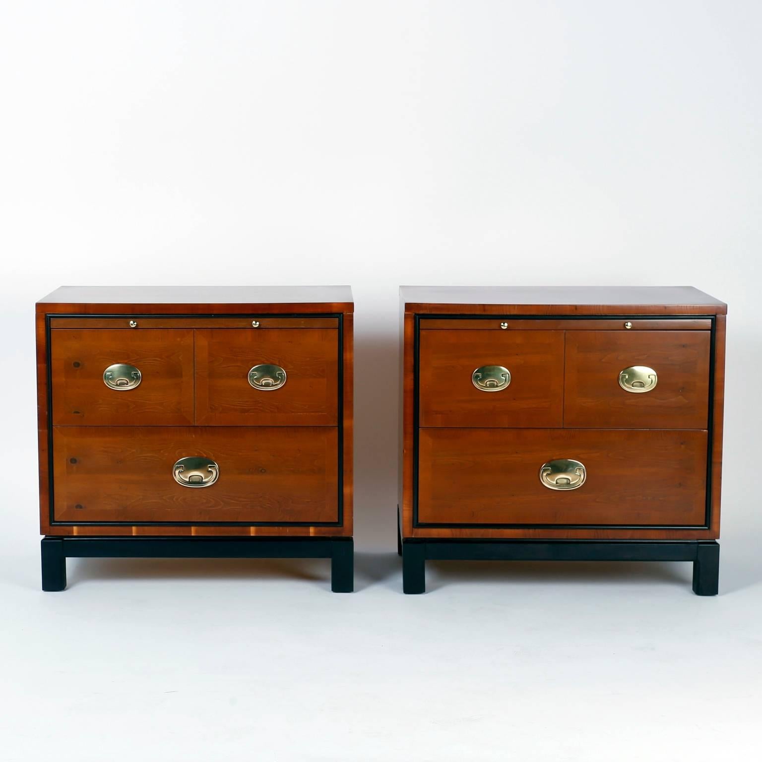 Pair of nightstands or three-drawer chests beautifully crafted with well grained walnut in a clean modern form. Featuring a pull-out tray, stylized campaign hardware and an ebonized Ming style base. Signed Hickory Manufacturing Co. in a drawer.