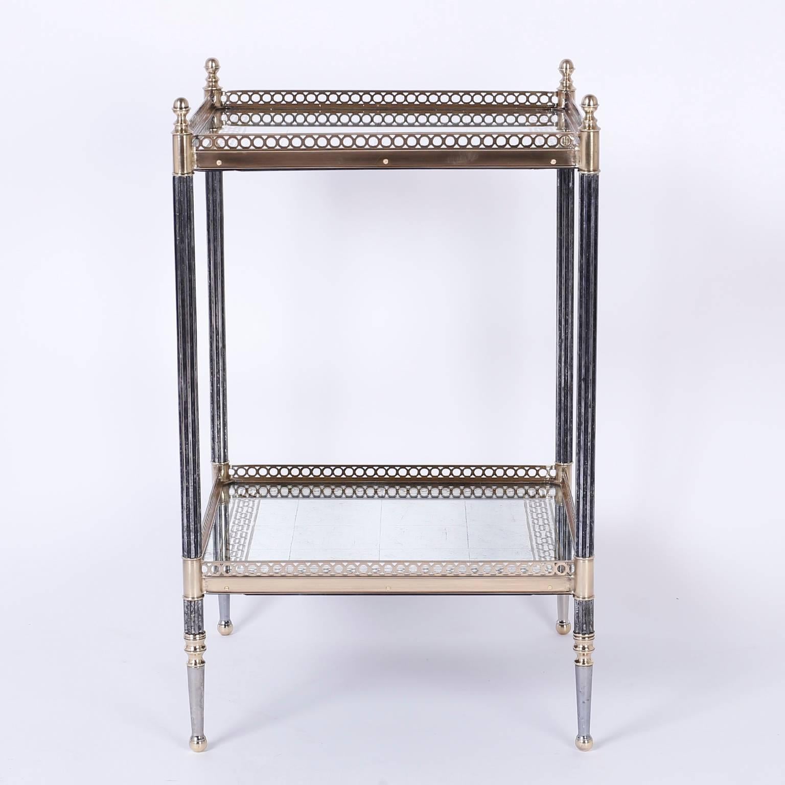 Two-tiered metal and glass table or stand with an elegant metal frame having finials at the top and ball feet. Each tier is silver and gold leaf under glass bordered by a brass gallery.