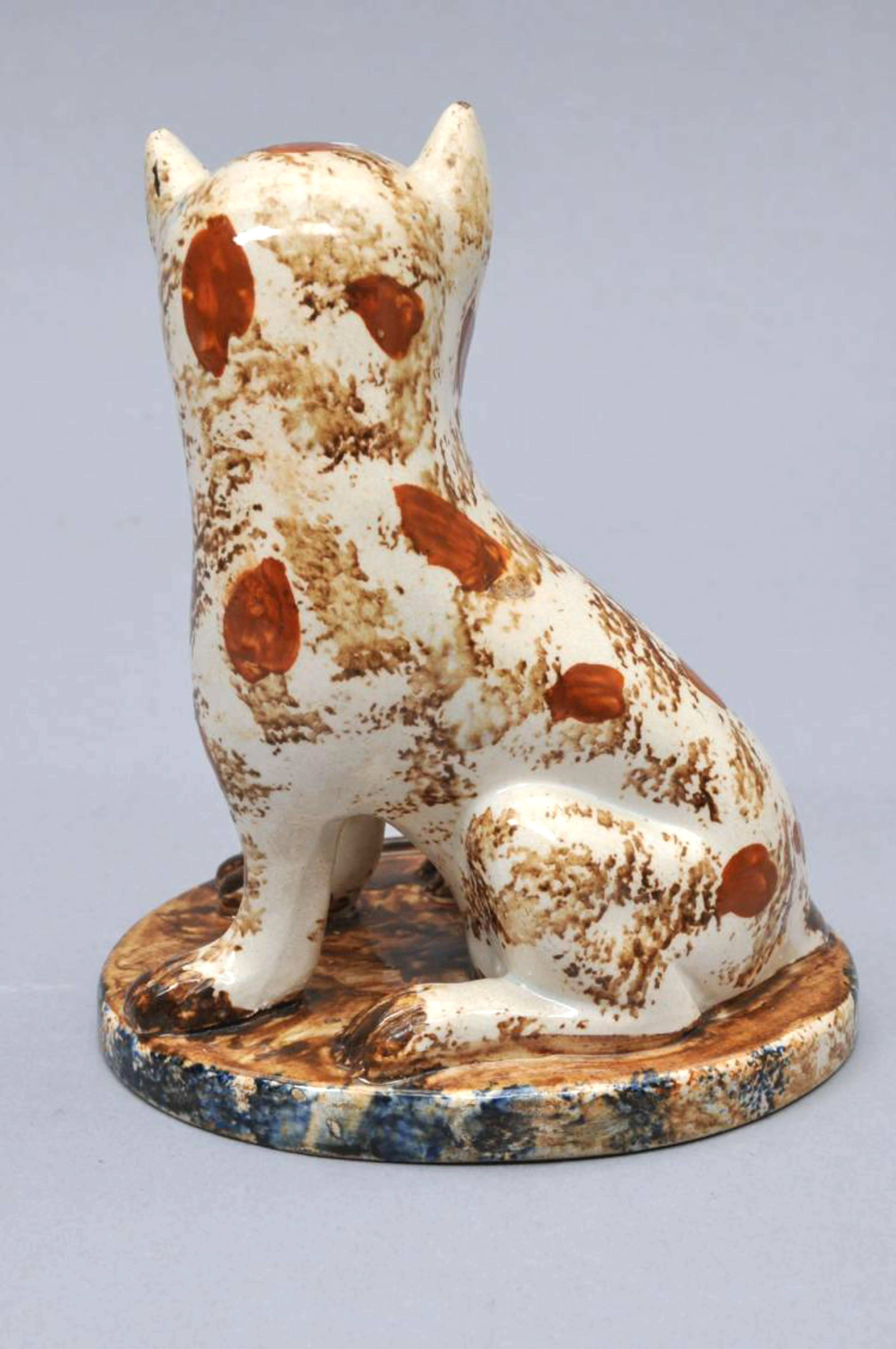 Early Staffordshire Pottery Cat, 
Early 19th century.

The large whimsical cat with brown and iron-red sponging sits on its hind quarters on an oval base with blue sponging with a bemused expression on its face. 

Dimensions: Height 5 1/2