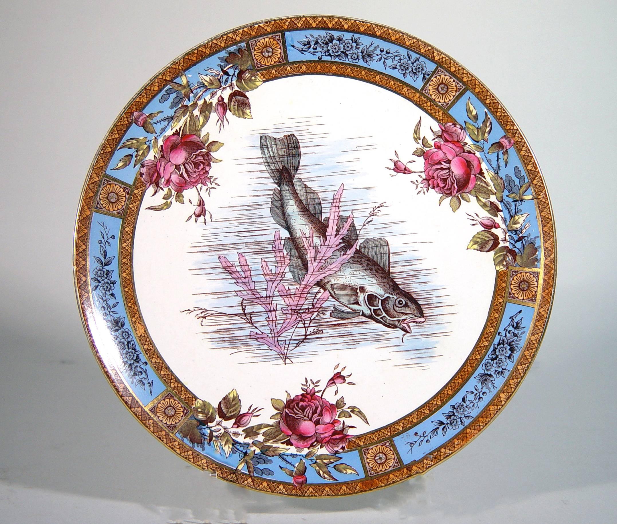 Les assiettes en poterie sont décorées d'un motif imprimé représentant un poisson au milieu d'algues dans une bordure de six panneaux bleu vif ornés d'une branche fleurie donnant naissance à des roses dans le motif central. Entre chaque panneau se
