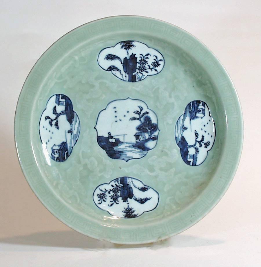 The Chinese porcelain circular dish has an everted rim with molded key motif and a shallow well molded with cloud scrolls.  The dish is glazed in celadon with five-lobed reserves decorated in underglaze-blue.  The central reserve depicts a figure