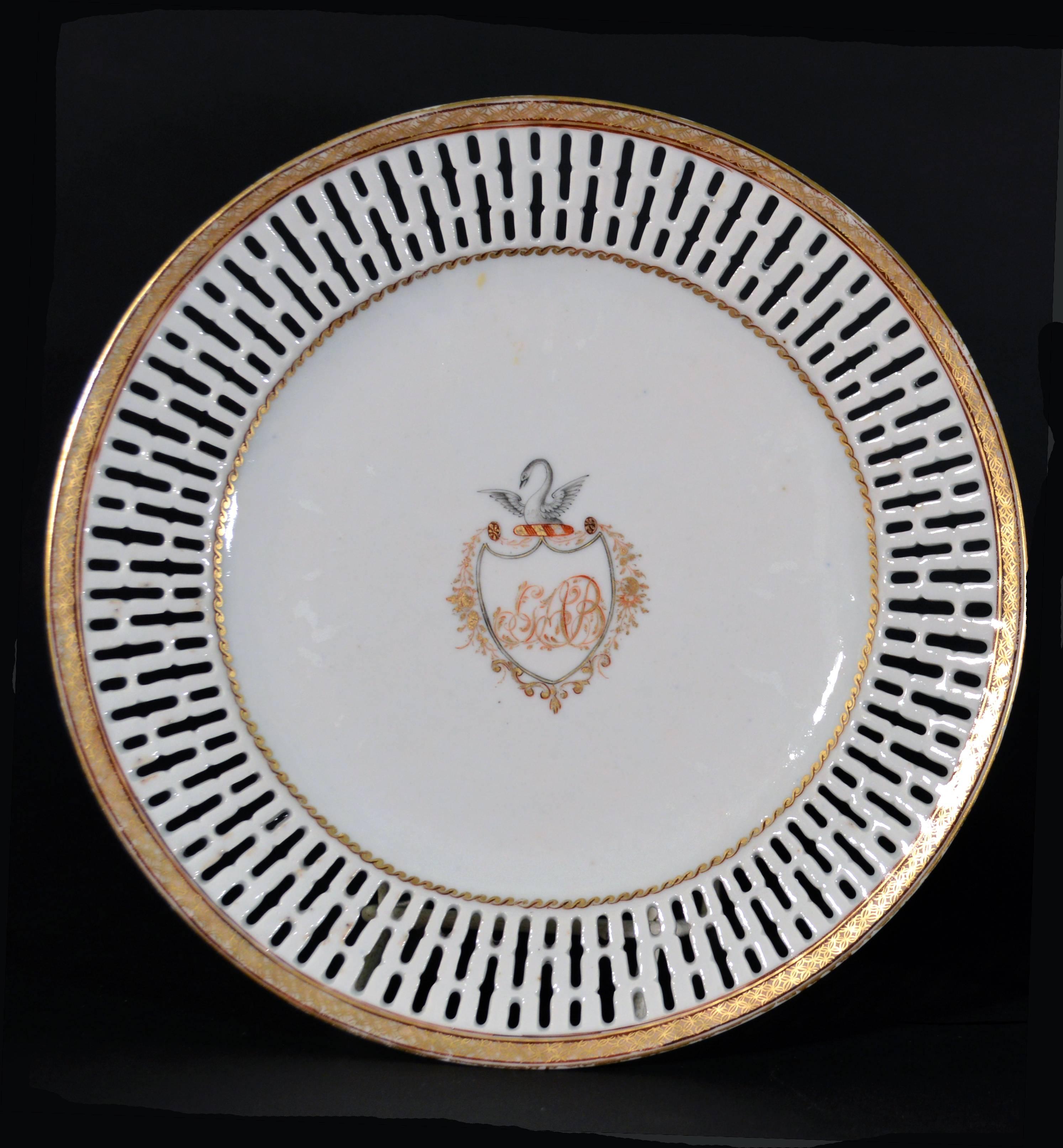 The plates are circular with an openwork inner border. The other rim decorated with tiny tight gilt flowerheads.  The outside of the central well decorated with a gilt and red ribbon.  

The center of the plate decorated above with a crest of a