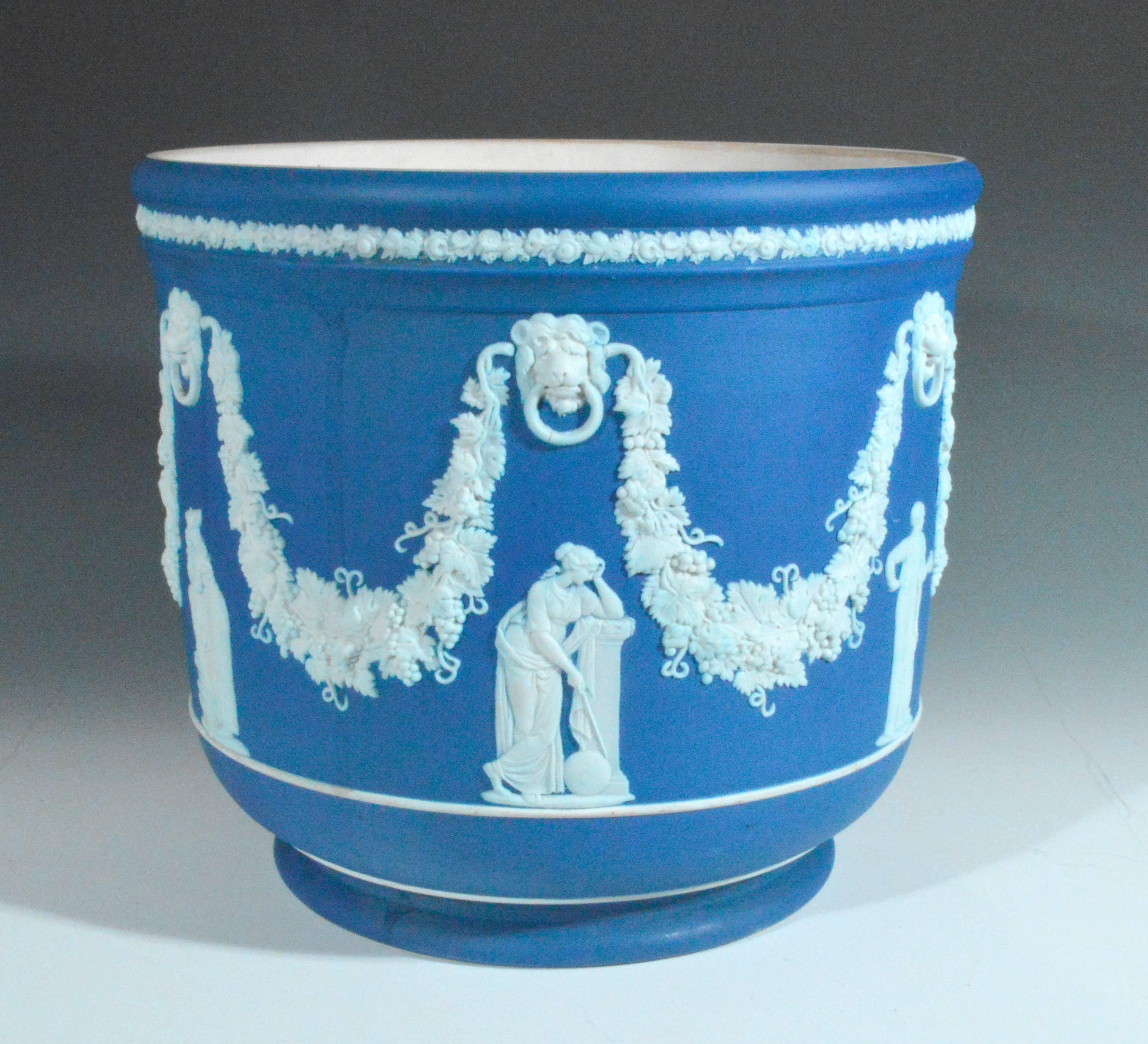The large pot is unglazed on the interior. The pot is decorated in a blue jasper with applied figures to six sides. Above each figure is an applied white lion with a vine leaf swag issuing from behind his head with large leaves and bunches of