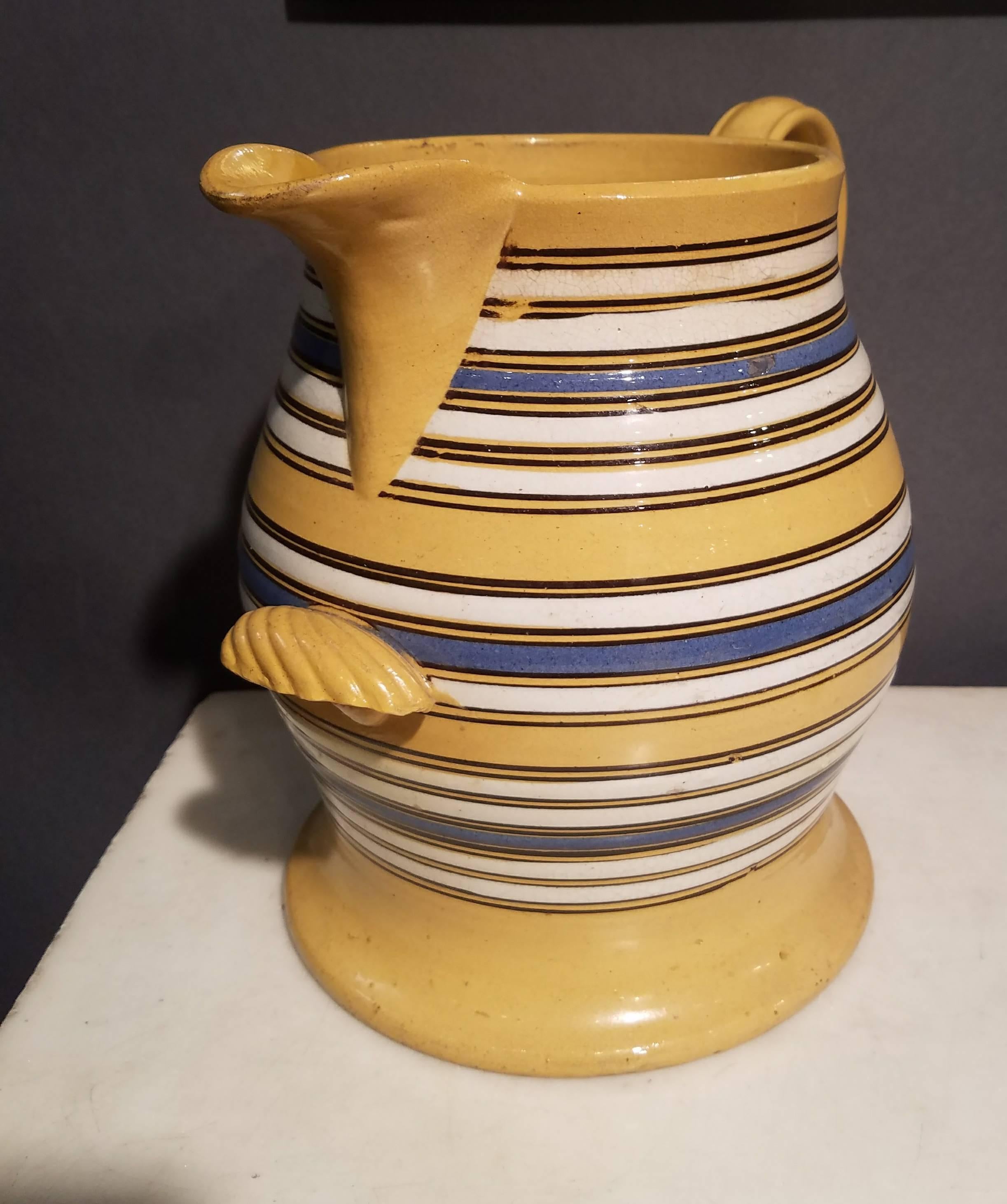Mochaware multi-banded large yellowware jug, 
circa 1840.

A dramatic large Mochaware yellowware jug with flared foot and multiple bands of solid colour in white blue and brown. Below the spout is a large sea shell grip (off/on) to help steady