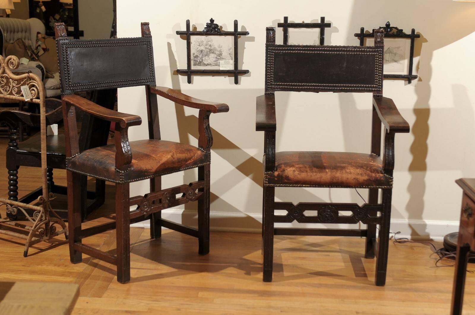 This pair of leather and wood chairs are Jacobean in style. 
The seats are leather and trimmed in nailheads. There are unique carvings on the legs, arms and backs.