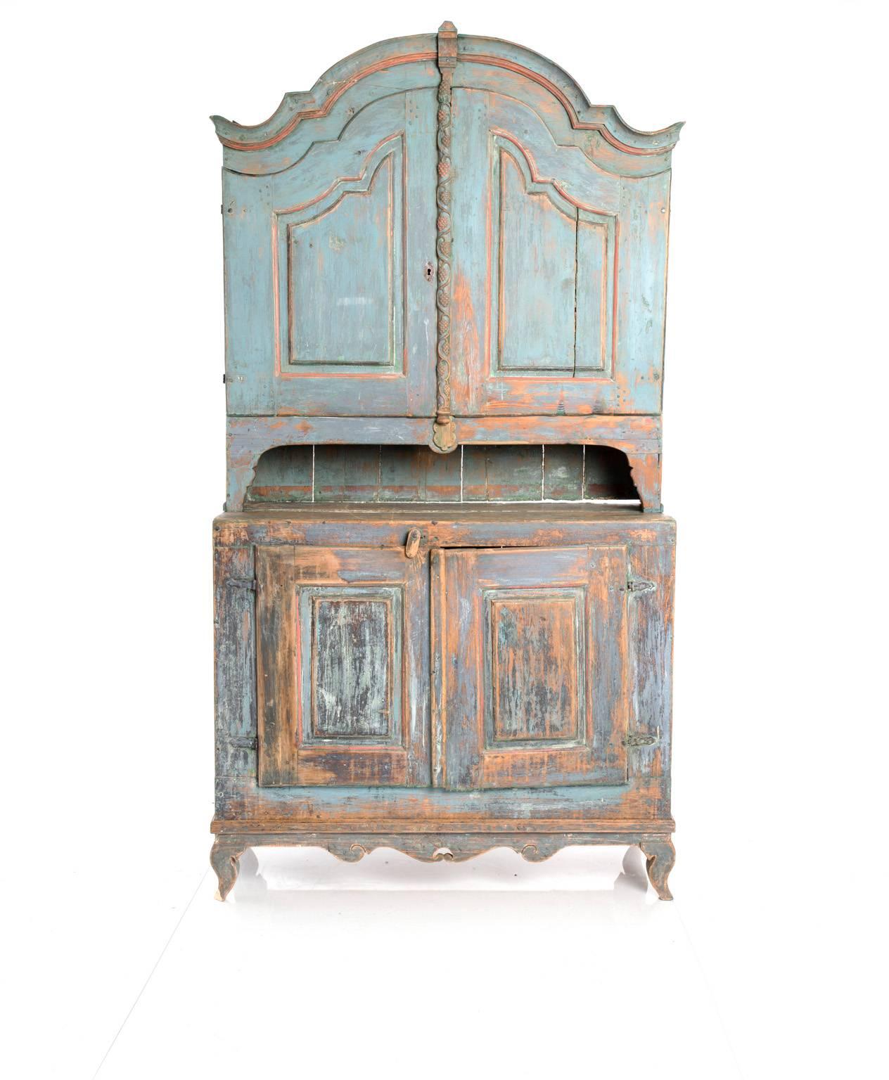 Sweden, circa 1780s. A blue-painted provincial Rococo-style cabinet. Upper case with rounded pediment, two doors and interior shelving. Lower case with carved skirt, two doors and interior shelving. Upper case doors have a border with carved grape