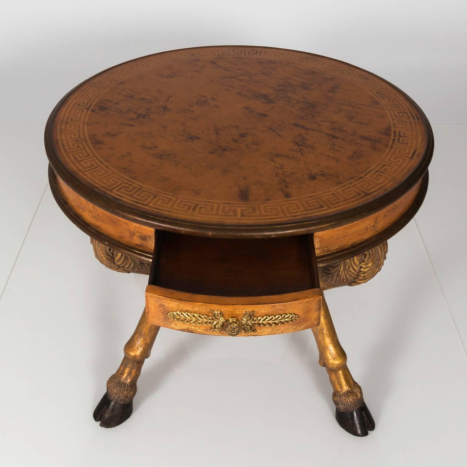 Early 20th century Empire style centre table resting on a goat shaped four legged base with round leather top and Greek key banding. The table skirt is leather with four small drawers embellished with brass mounts. Legs are hand-carved gilded and