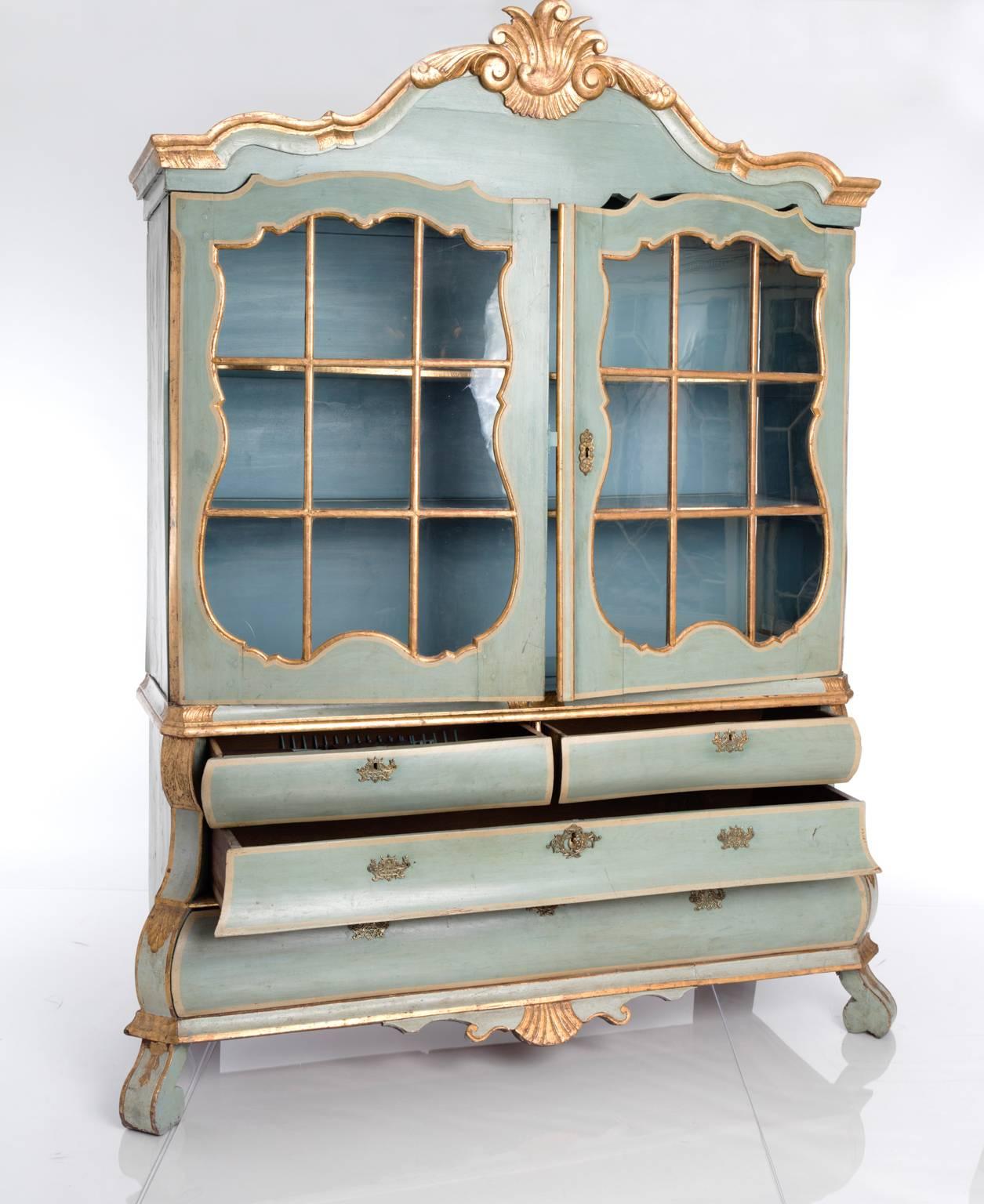 A blue-painted and partially gilt glass-fronted display cabinet with carved detailing, upper half with two glass doors and interior shelving, lower half with four drawers. Holland, late 18th century.
 