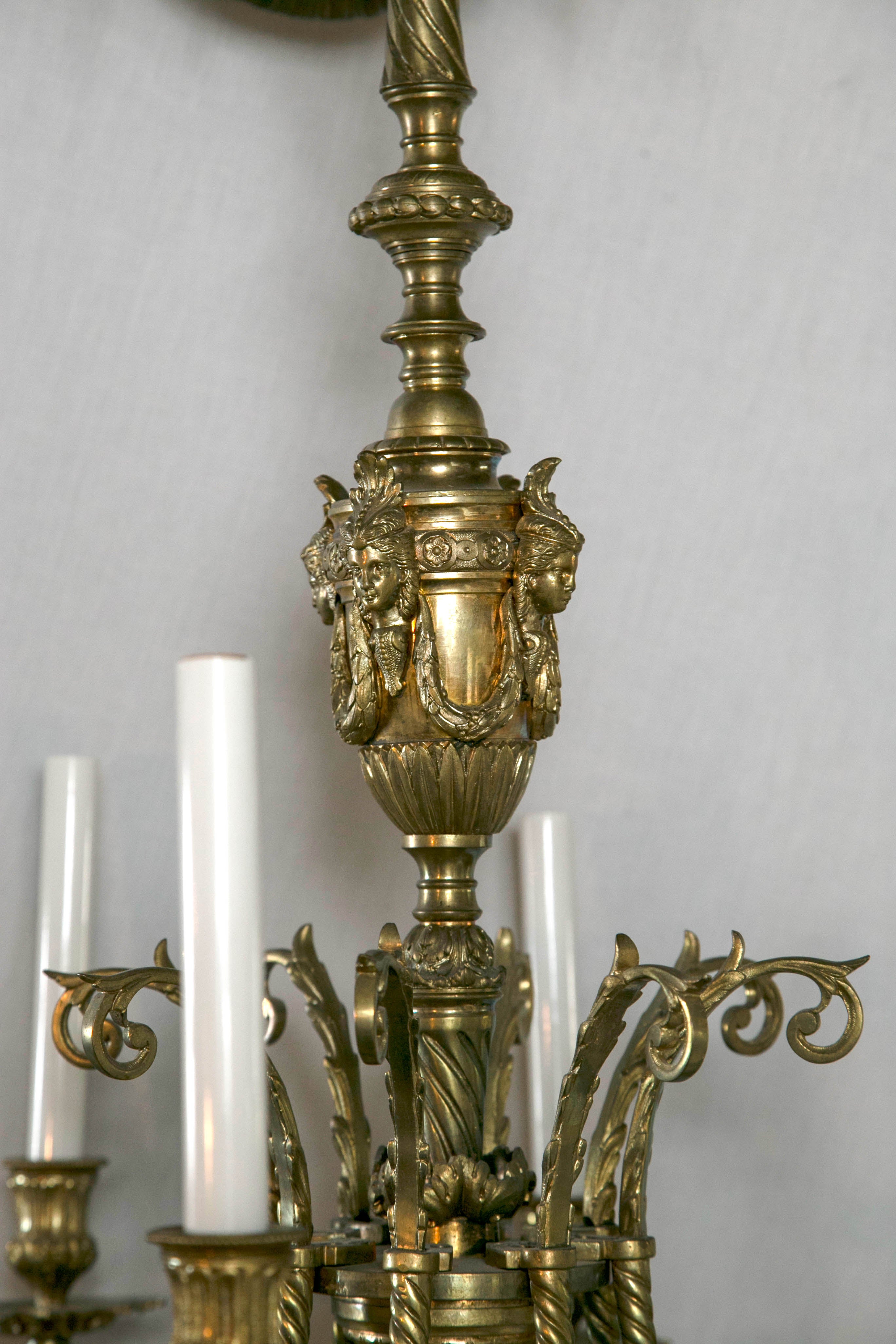 At the top is a typical  Louis  XVI style ribbon, below which is a  round  leaf and cluster form above the central column. Lady's  heads joined by swags above the  central part of the column to which are attached  8  arms decorated with leaves and