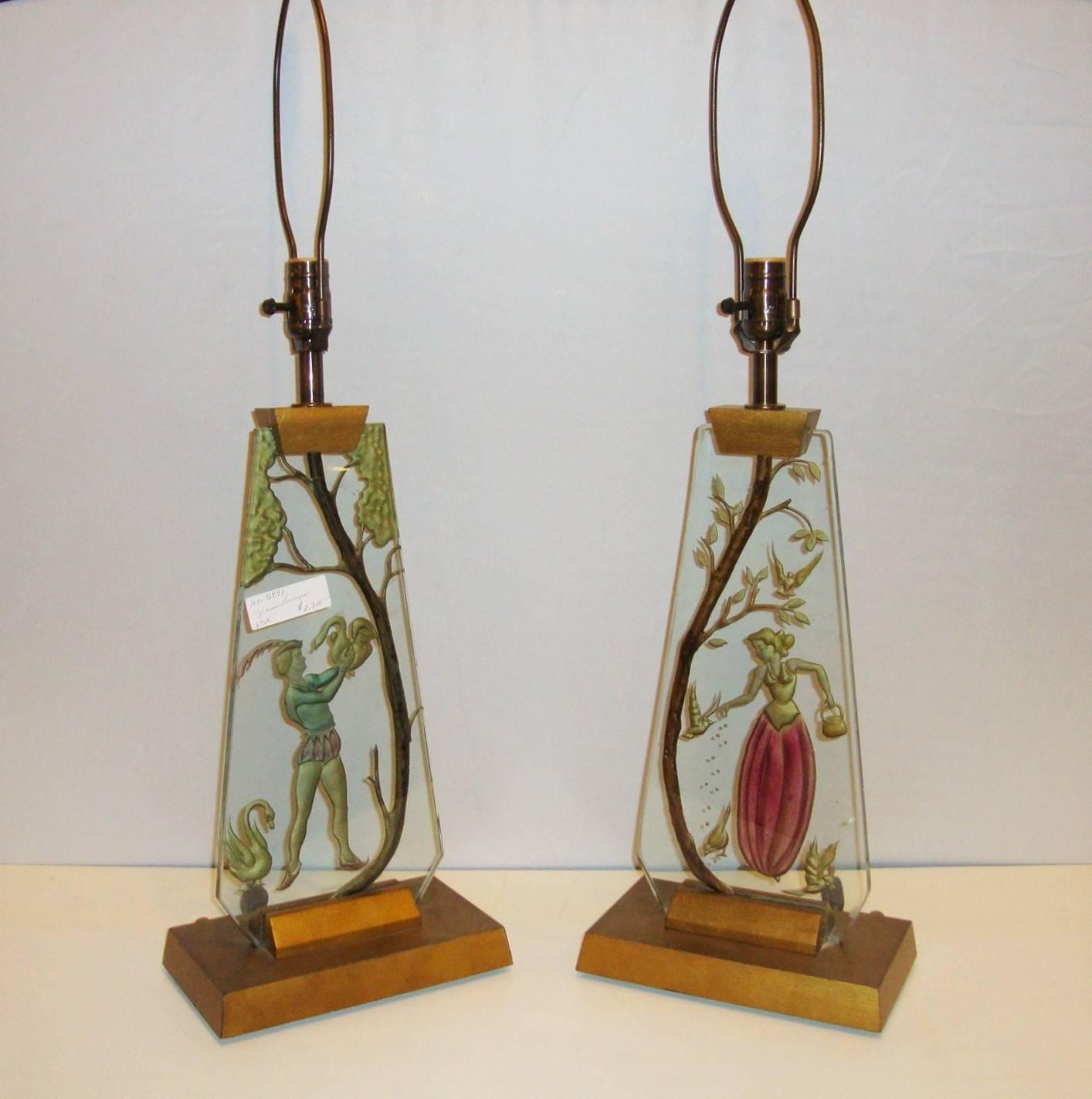 A pair of glass Mid-Century Modern table lamps. This elegant pair of Italian reverse painted glass lamps detail one hand-painted man enjoying some geese, while the other shows a woman graciously feeding birds. Both on wooden bases and wooden tops.