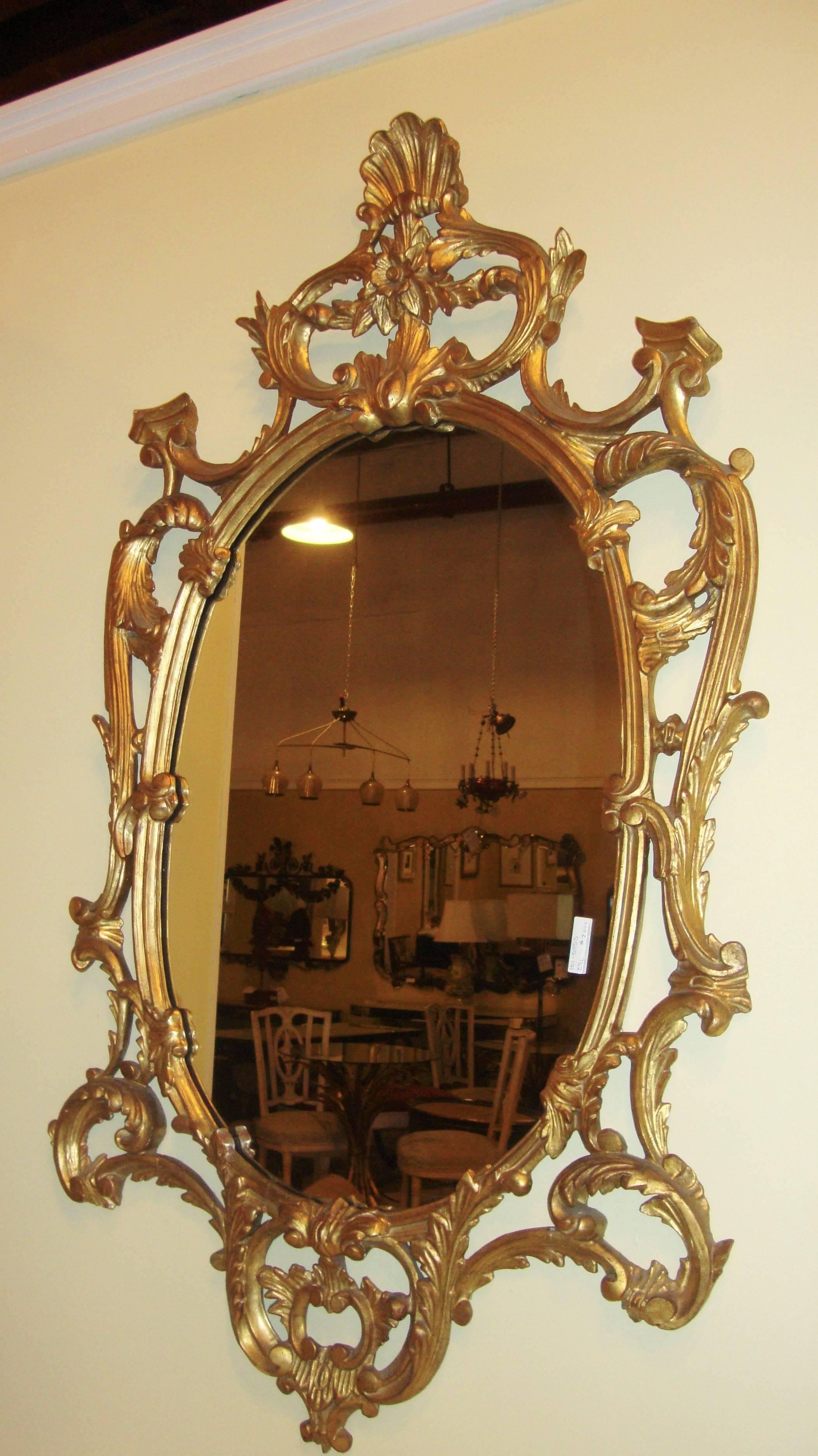 An Italian oval giltwood framed wall mirror. The center mirror having a giltwood frame from the 1960s of Italian origin. The overall theme of the frame is carved scroll and leaf with vine work which terminates in a rose and shell carved pediment.