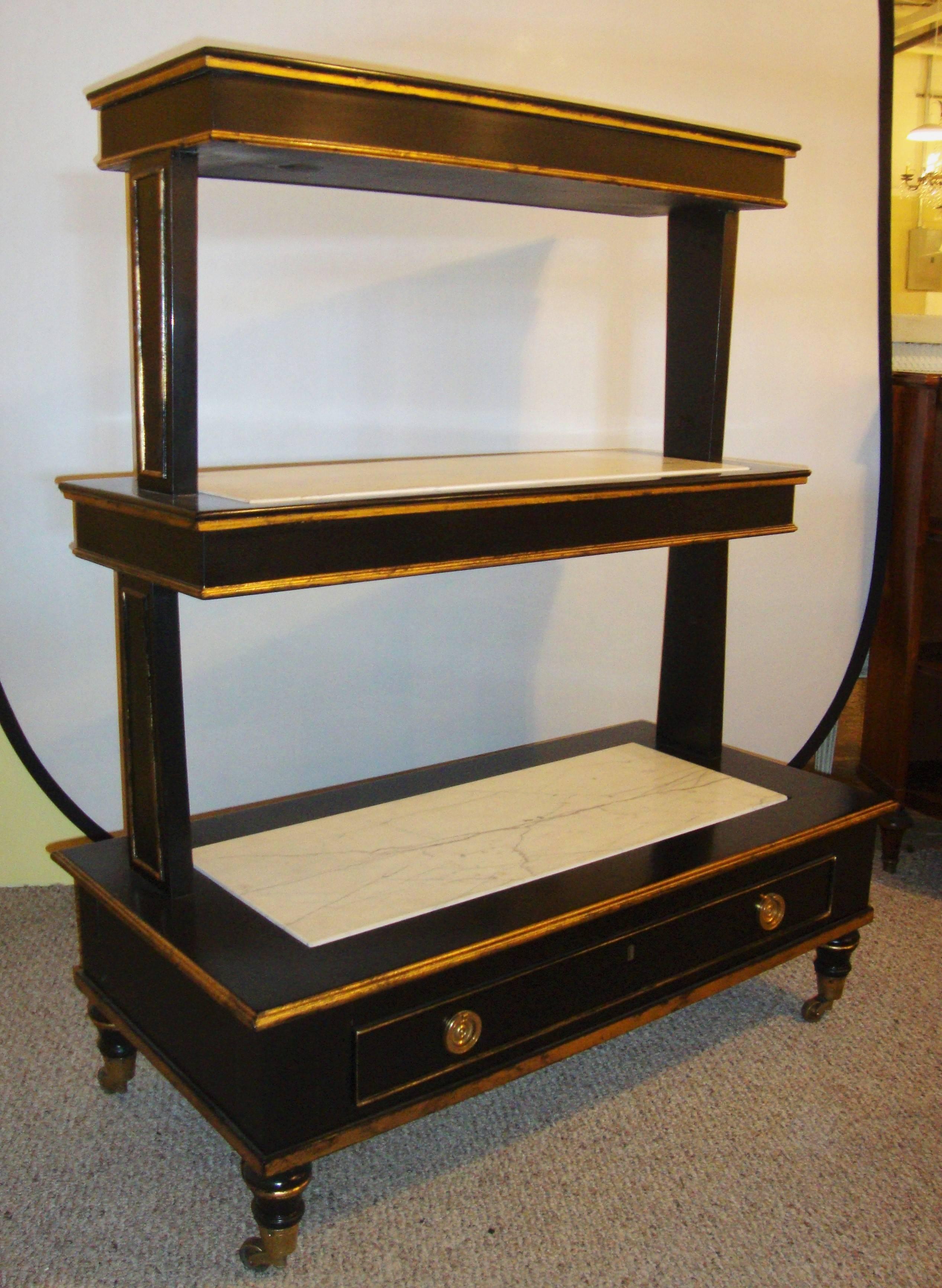 An ebonized three-tier bookshelve with a single drawer below. Marble tops on each of the shelves, gilt highlights throughout on rolling wheels.