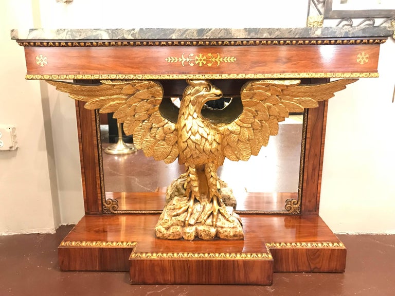 A pair of Regency style brass inlaid rosewood parcel-gilt and marble consoles. Each green marble rectangular top above an egg-and-dart-carved edge supported on an outspread winged eagle support with mirrored back, on a break fronted plinth.