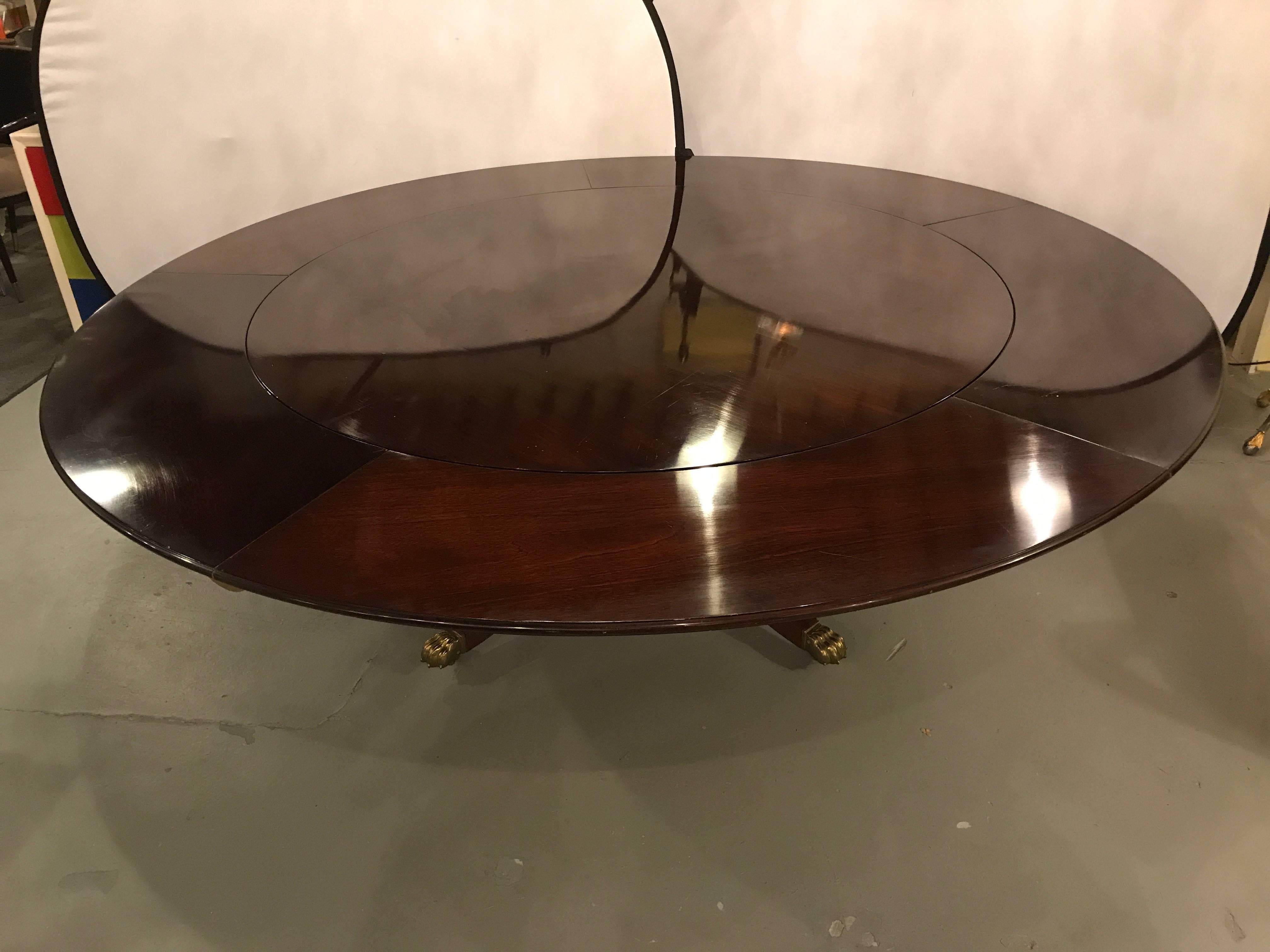A circular dining table by William Tillman. This fine solid mahogany dining room table is made of flame mahogany having five 14 inch interlocking leaves and sits atop a quad based pedestal with a lower shelving area on sprayed legs terminating in