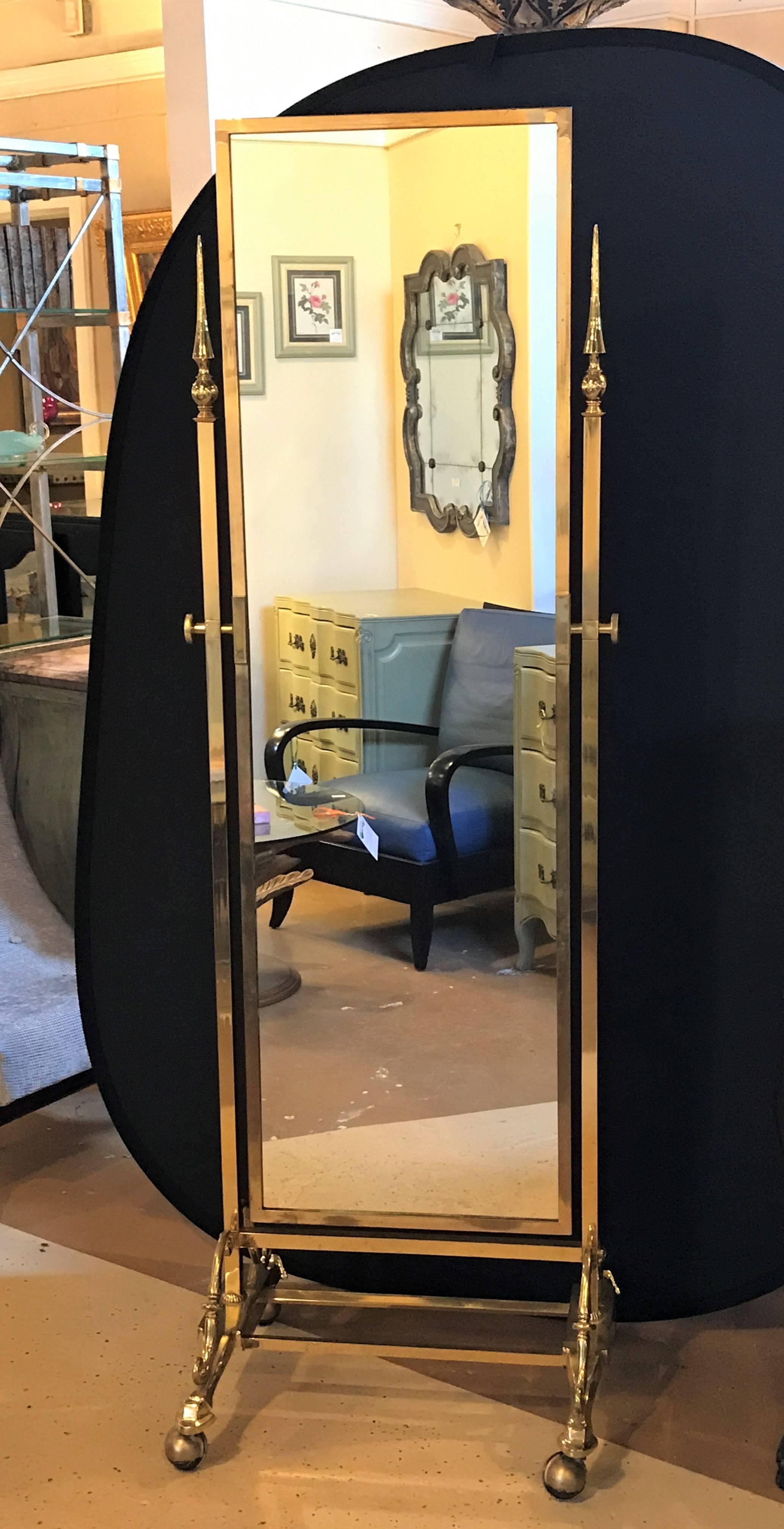 Hollywood Regency style brass cheval mirror. The whole on rolling caster feet leading to a rectangular pole column-form side terminating in a pointed finial supporting a brass framed center mirror.