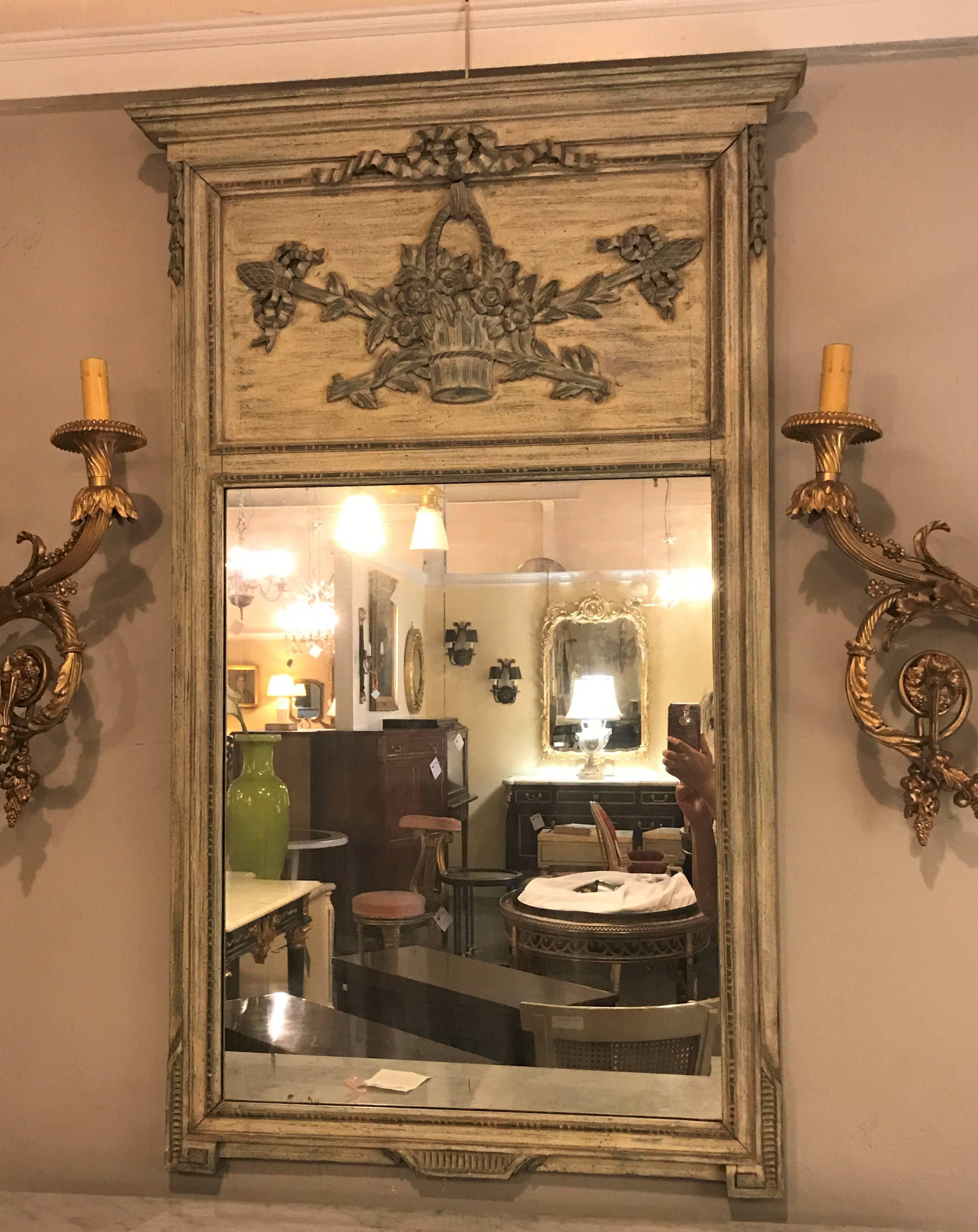 A French Trumeau beveled wall mirror. Paint Decorated having a wooden back and uniquely carved top depicting a basket of flowers with crossing swords. In a distressed finish.