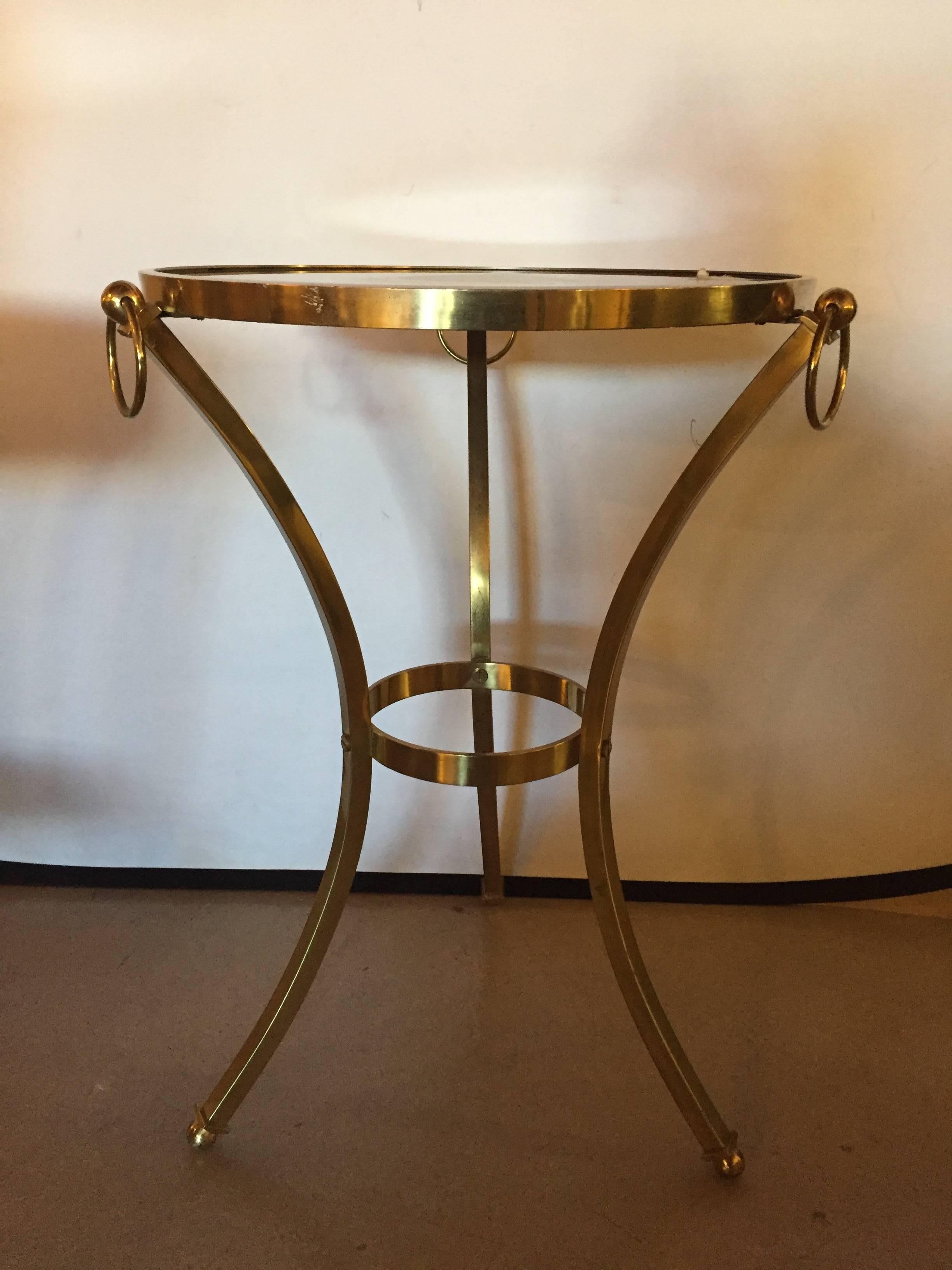Small brass Hollywood Regency gueridon table with glass top. Typical in form and style comes this inexpensively priced end or gueridon table having an ebony glass top supported by a brass apron having ti-pod stand with circular form on the top and