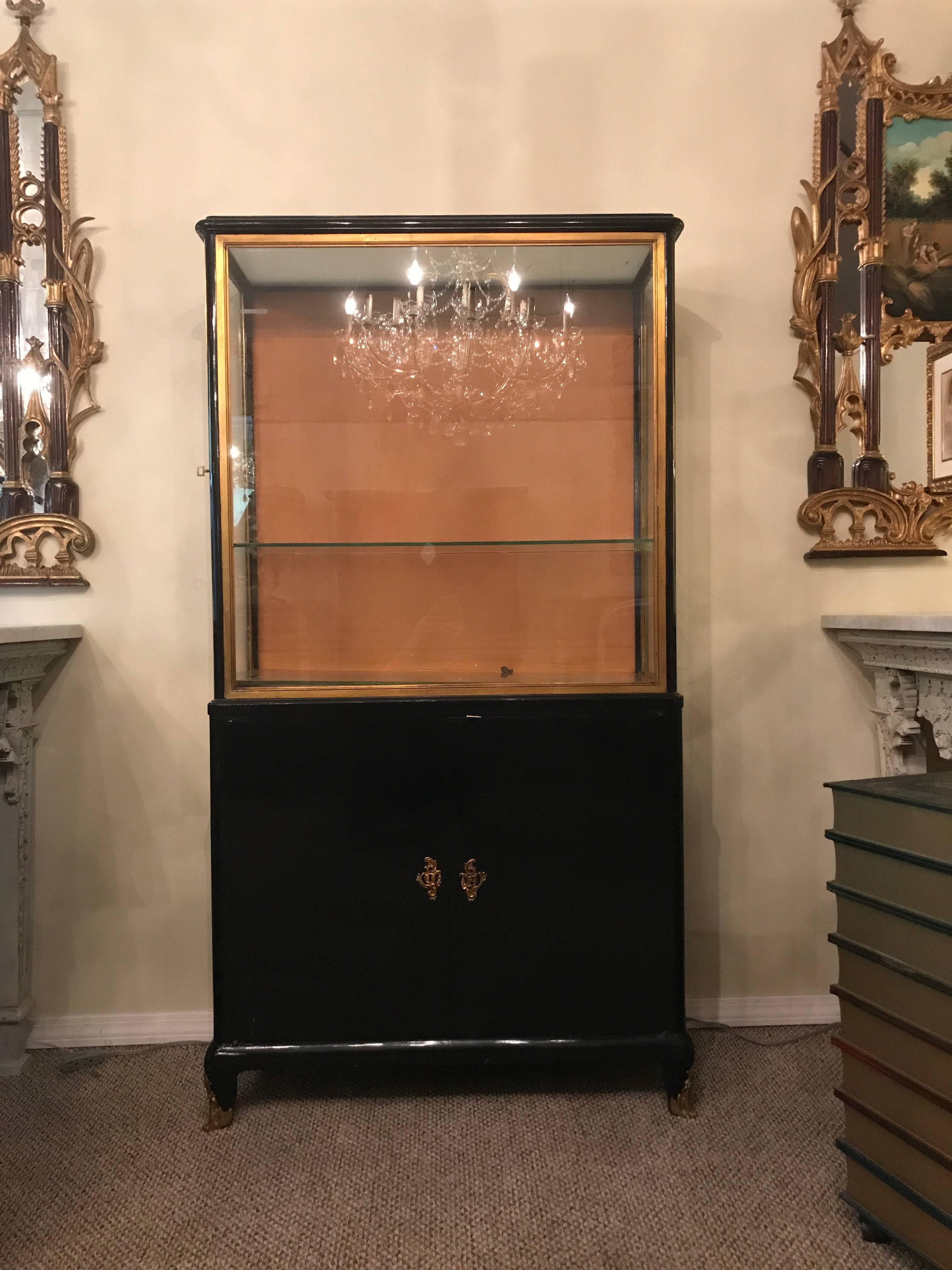 Ebonized vitrine or China cabinet bookcase manner of Jansen. The bronze sabot feet supporting a pair of lower doors under a showcase top in a bronze frame which opens from the sides rather than the front. Lighted.