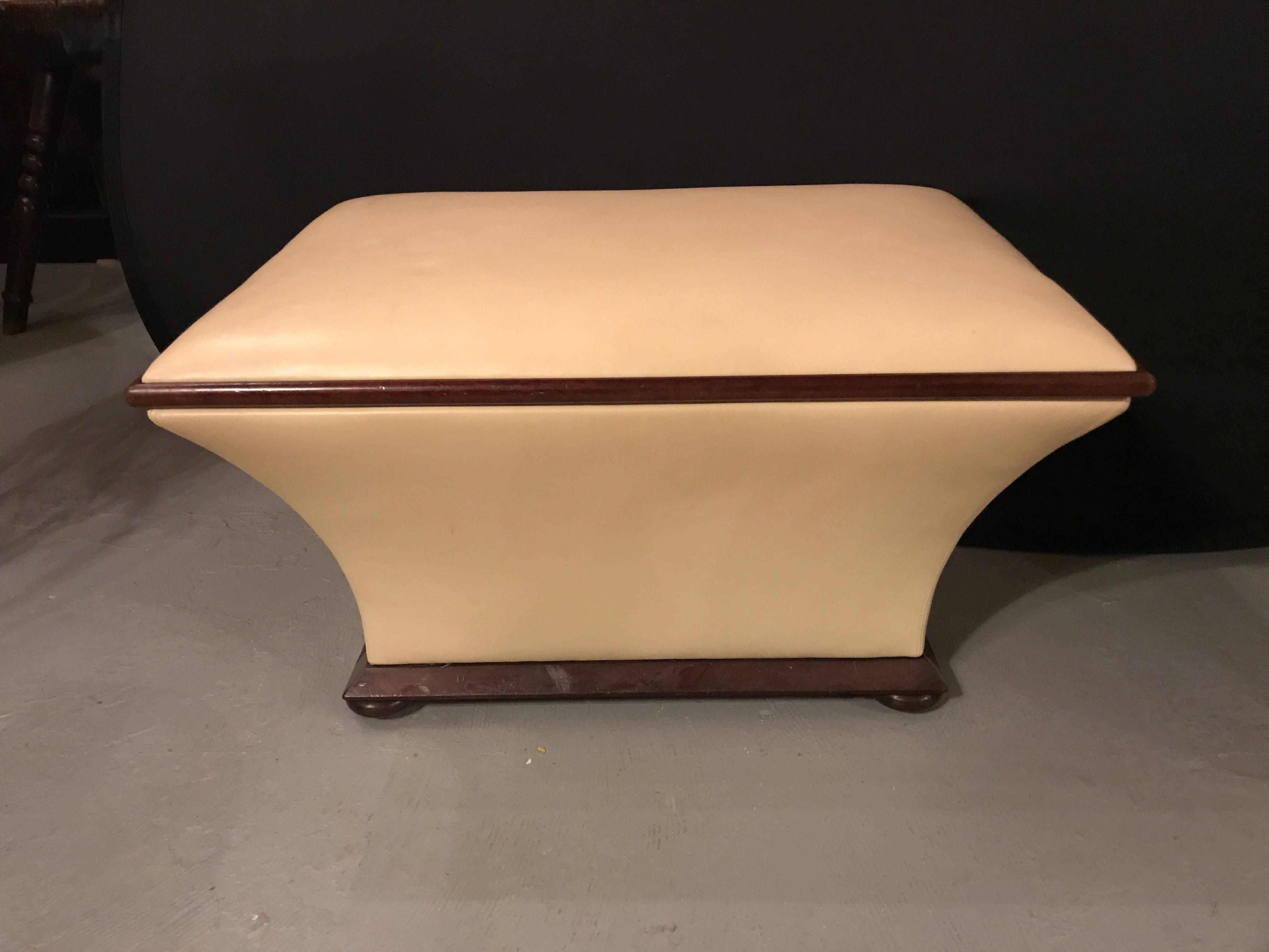 A Hollywood Regency style leather stool chest. Having sleek and defined lines this curved bench or footstool is comprised of mahogany trim and sits on bun feet. The tan leather frame has a pull up top with straps and leads to a large open interior