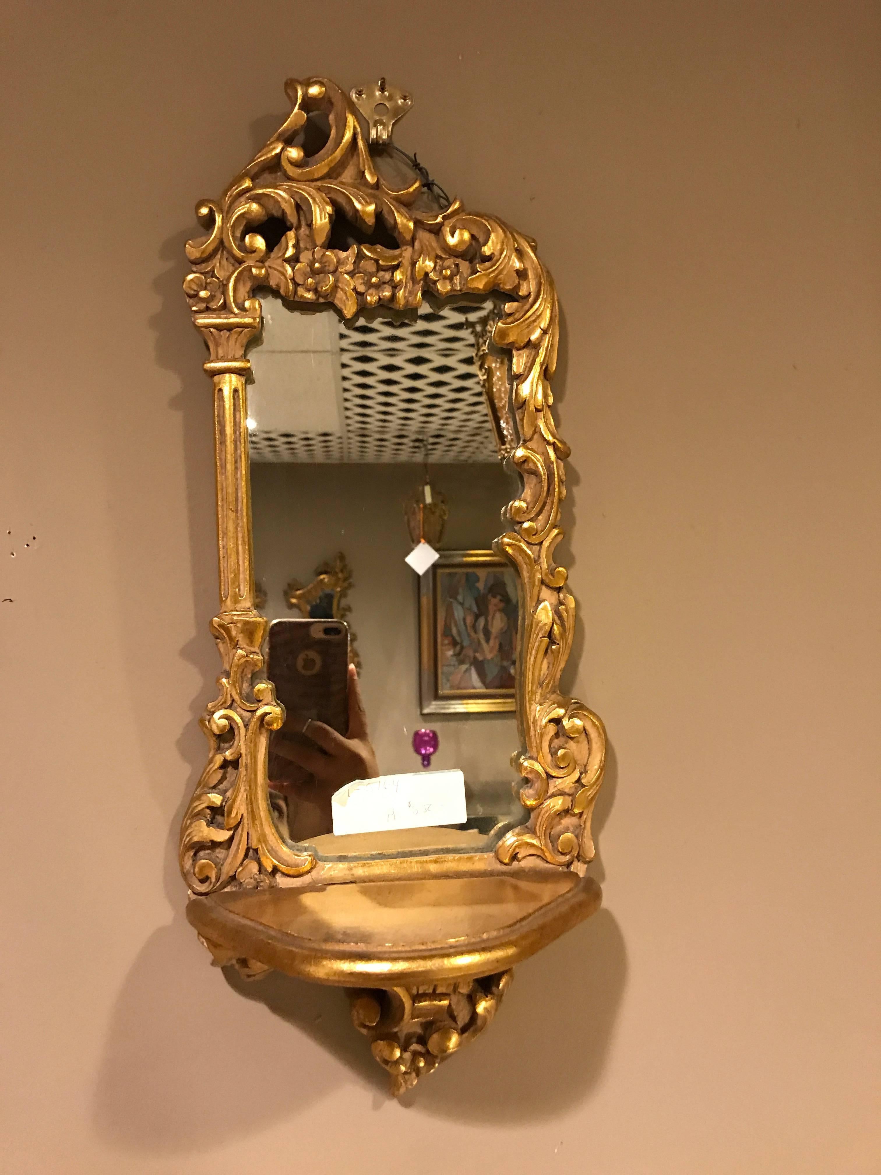 Pair of diminutive giltwood mirror back wall shelves. The giltwood frame having a single shelf for small nick knacks. Each having opposing column-form and carved sides and floral and leaf designed carved tops.