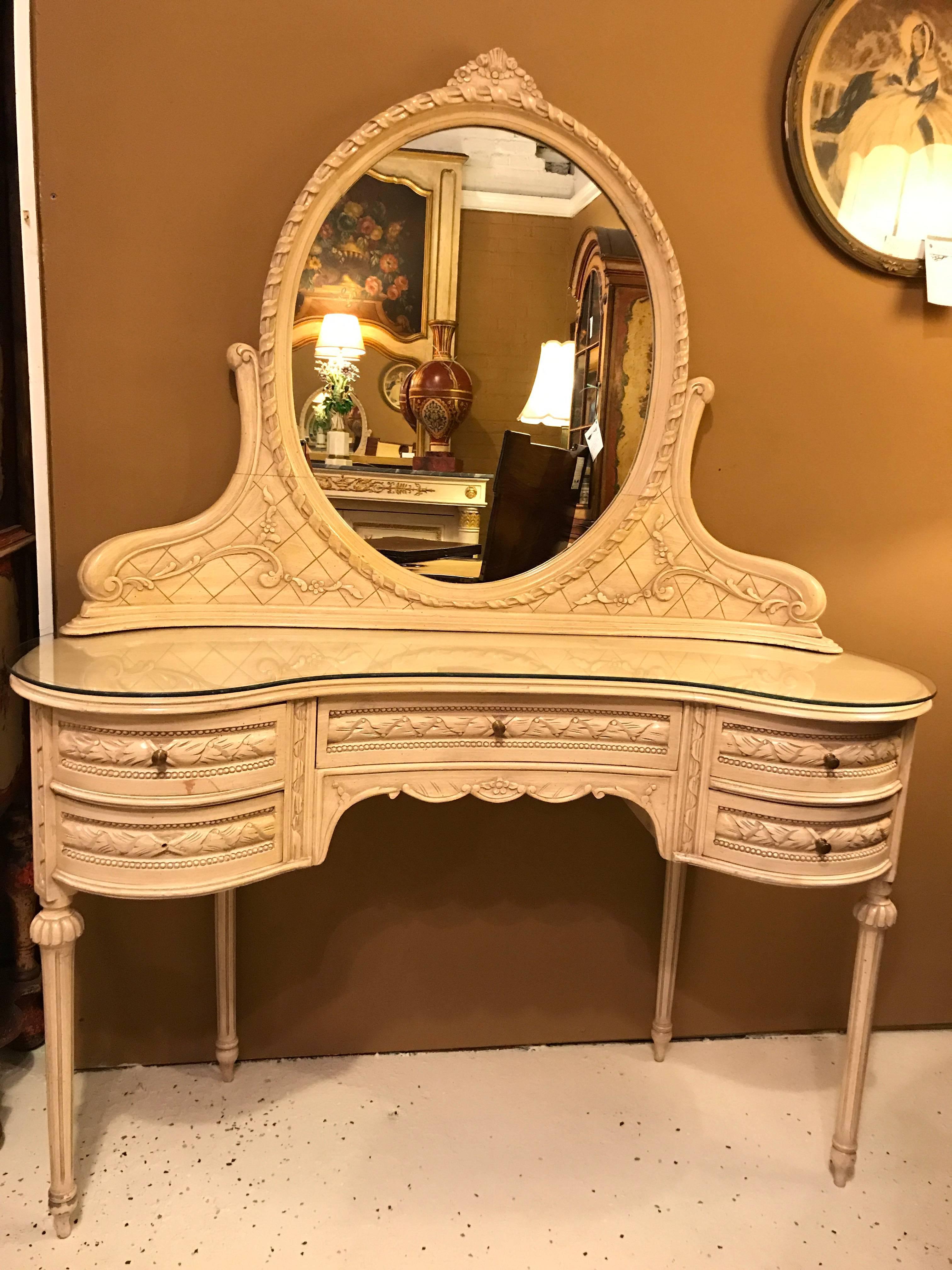 A Hollywood Regency vanity desk with mirror and chair in Swedish Fashion. This Louis XVI style vanity desk has five drawers and an oval detachable mirror with a matching chair. The whole done in a distressed finish of off-white. A fitted glass top