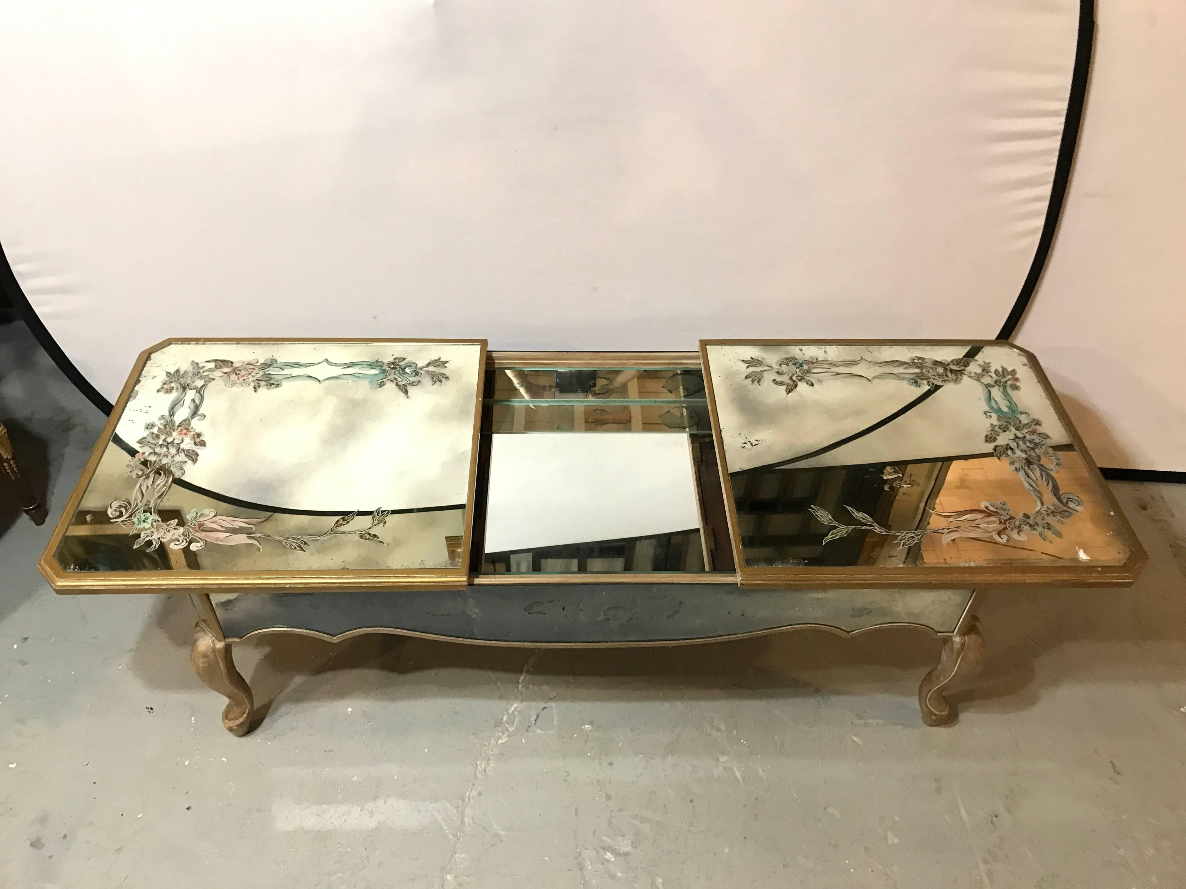 An Italian reverse paint decorated sliding top coffee low table. The gilt gold design legs and mirrored apron support a floral arrangement reverse painted on a mirrored top. The end slides open to use for storage.