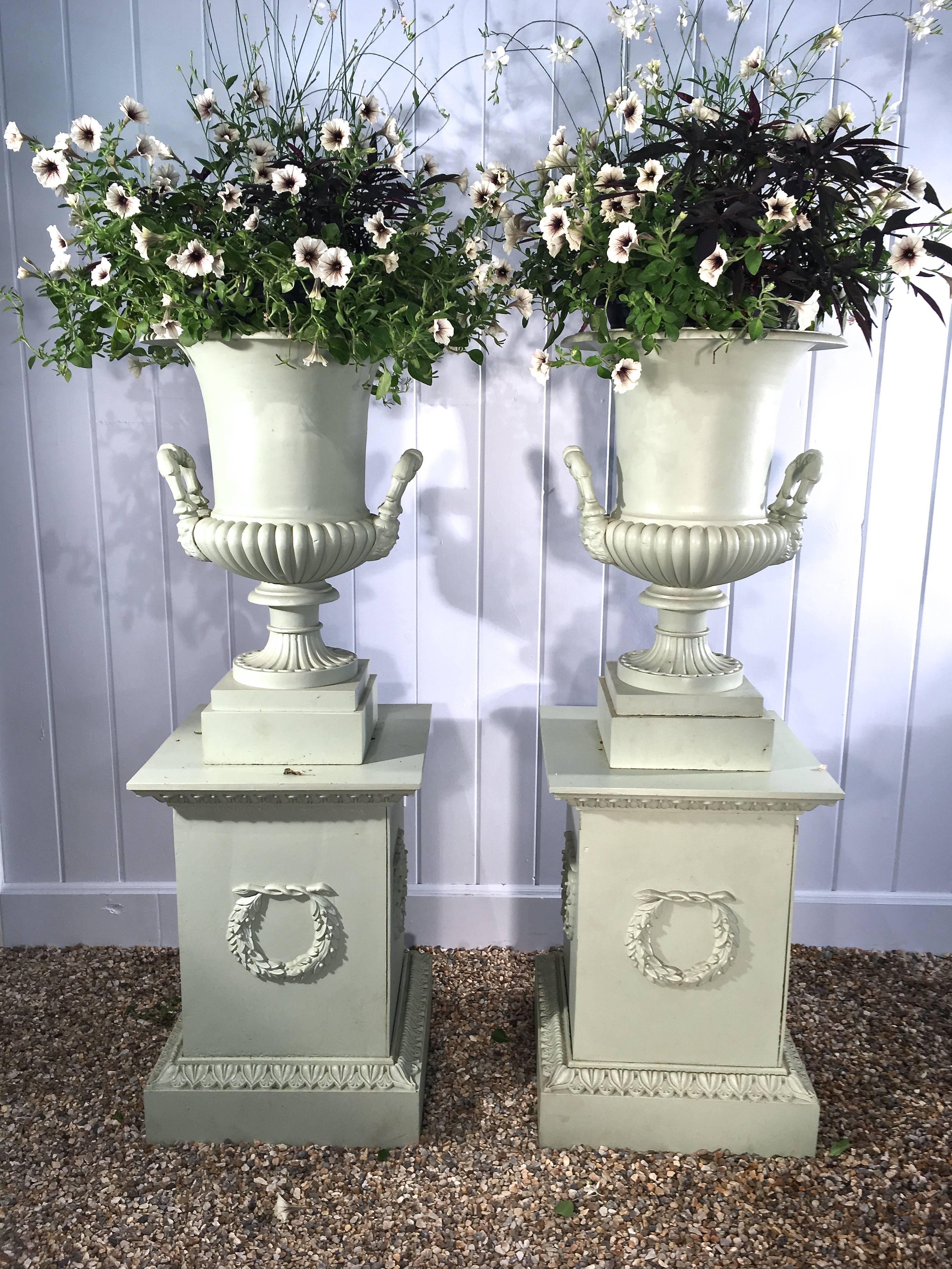 These are an outstanding pair of cast iron campana-form urns with plain everted rims, quarter-lobed bodies, and flared masked handles on reeded round feet. Surmounted on a conforming pair of cast iron plinths with egg and dart borders and wreathed