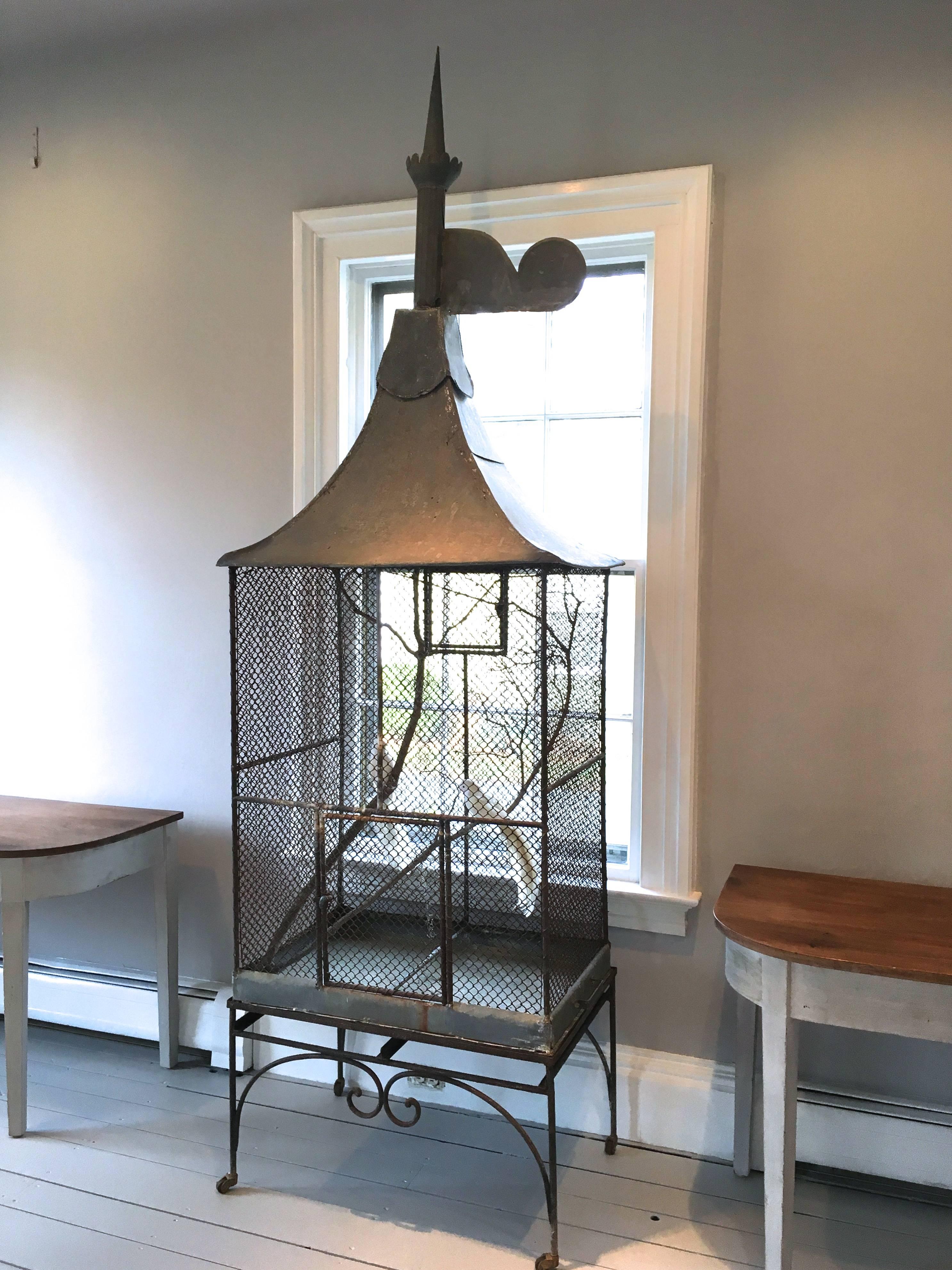 An elegant and unusual wrought iron parrot cage with zinc pagoda roof and grand finial sits snugly on it's original wrought iron stand with operational wheels. Original slide-out litter tray and two access doors make this piece perfectly functional
