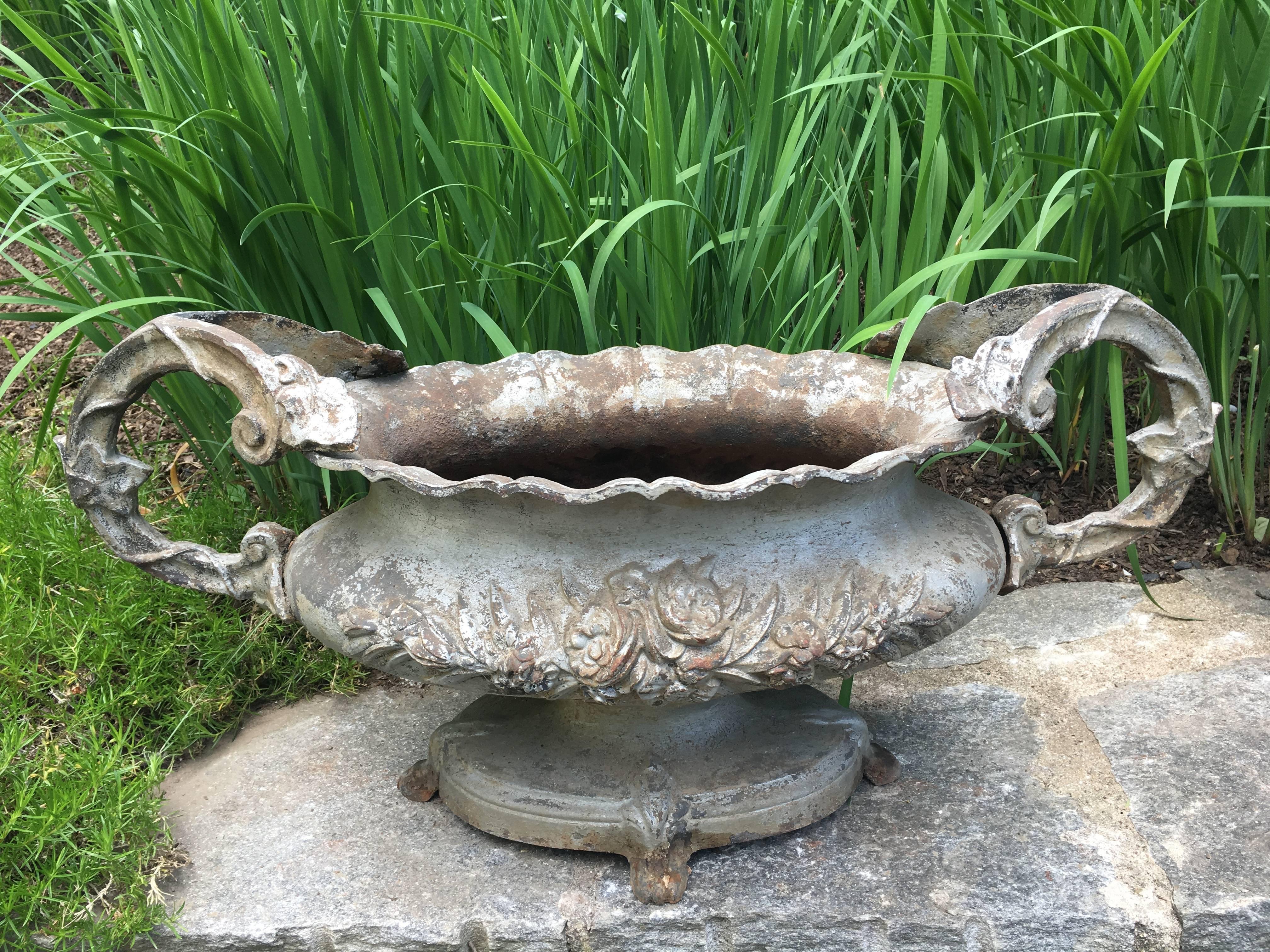 This large tabletop cast iron urn has crisp, detailed casting, elaborate handles, and an oval base with feet. In old silver paint with a bit of surface rusting, it is in wonderful condition with no chips, cracks, breaks, repairs, or missing bits. It
