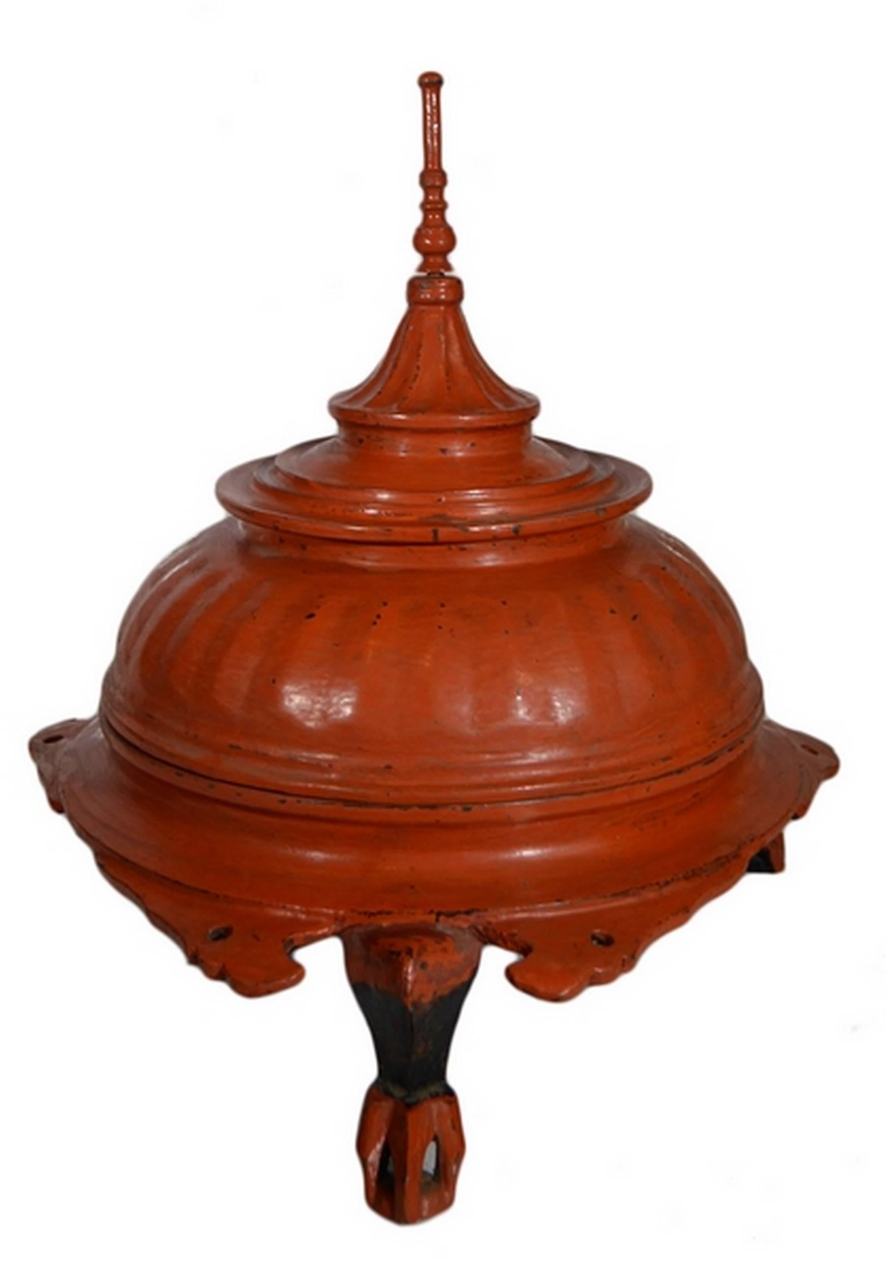 A 19th century Burmese offering basket made of carved wood painted red. This round basket displays a three-legged base with detailed carvings on the edge. The lid adopts a Burmese temple shape, with a sharp top surmounting a gadroon dome. The whole