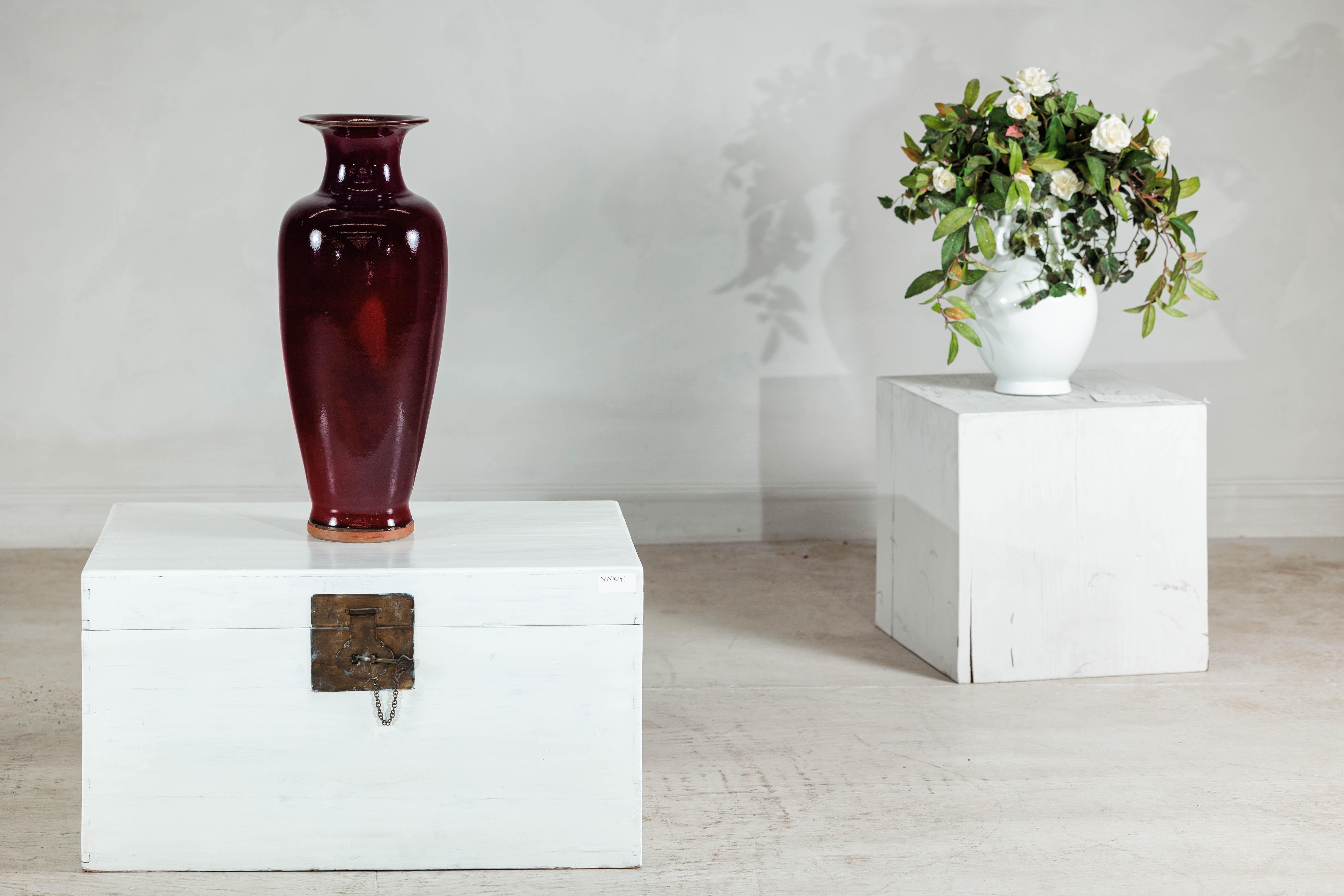 This tall vintage vase is distinguished by its richly layered oxblood flambé glaze — a deep burgundy red that subtly shifts toward violet and plum, especially near the rim and shoulders. Fired in a reduction atmosphere, the glaze creates moments of