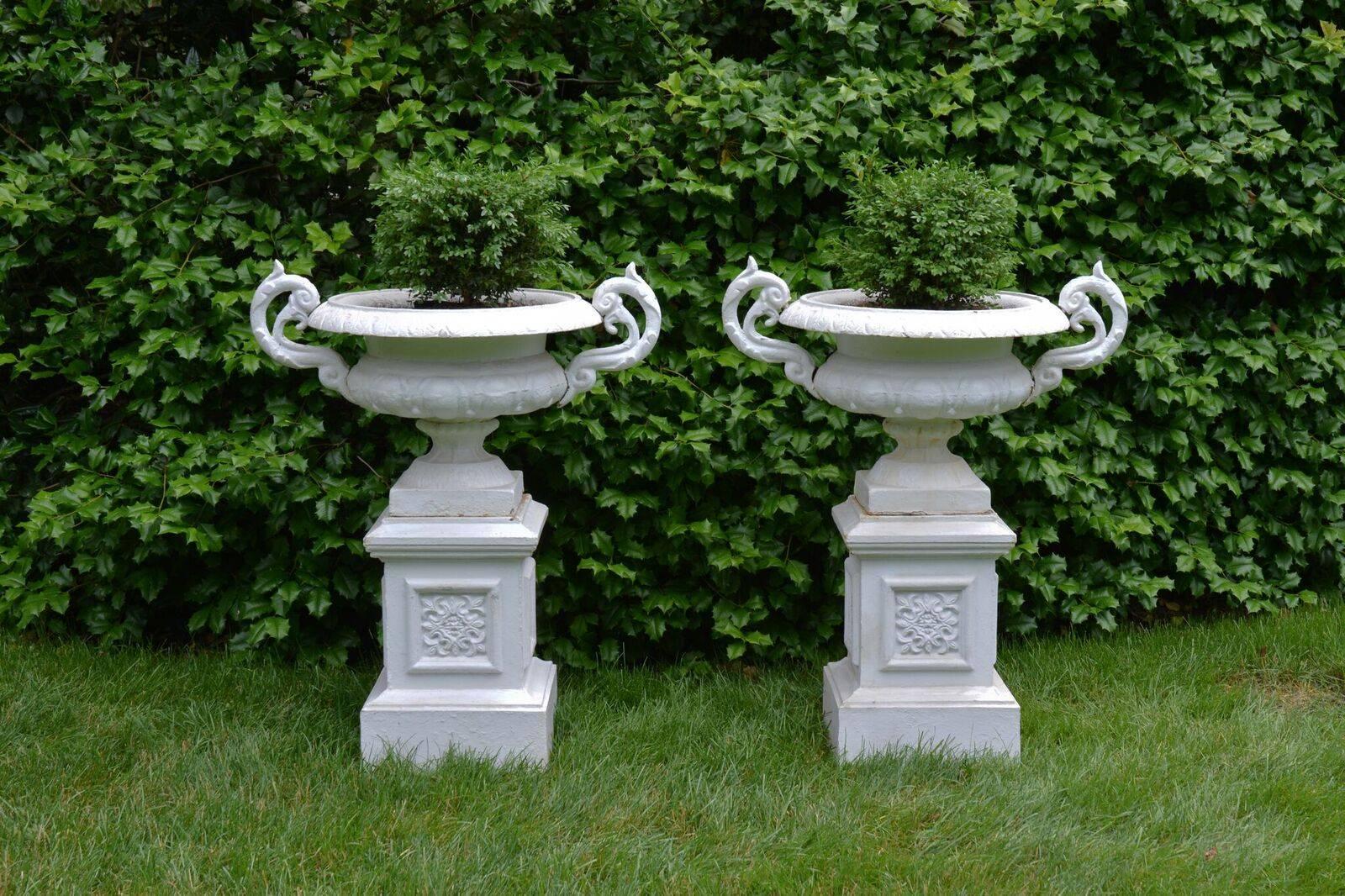 A pair of cast-iron tazza form urns with scrolled handles, the body of the urns semi-lobed and on rising socles with egg and dart motif, on integral square bases, the urns atop cast-iron pedestals, the side panels ornamented with stylized botanical