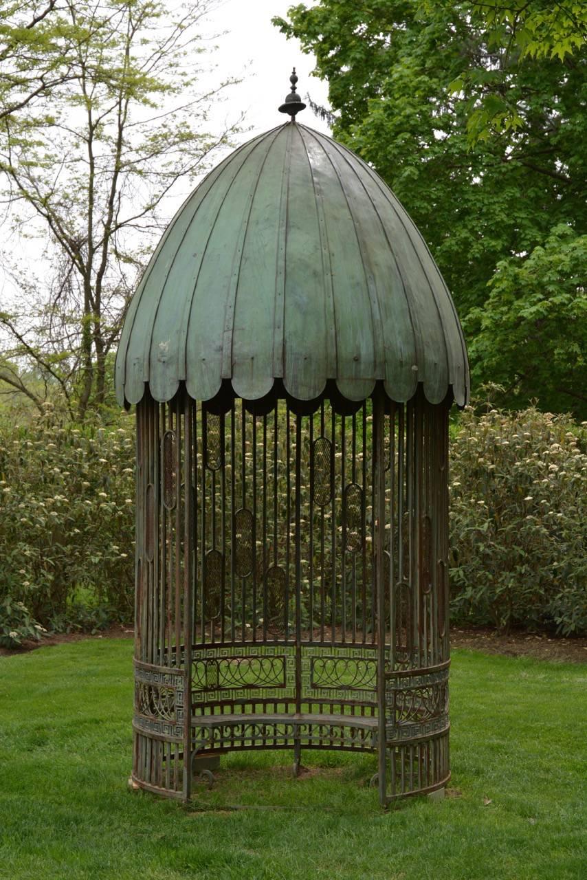 An unusual wrought iron garden pavilion with domed, scalloped copper sheet roof and vertical uprights above a wide band of looped iron and Greek key border with semi-circular interior bench, interior dome fitted with ancient-style oil lamp.