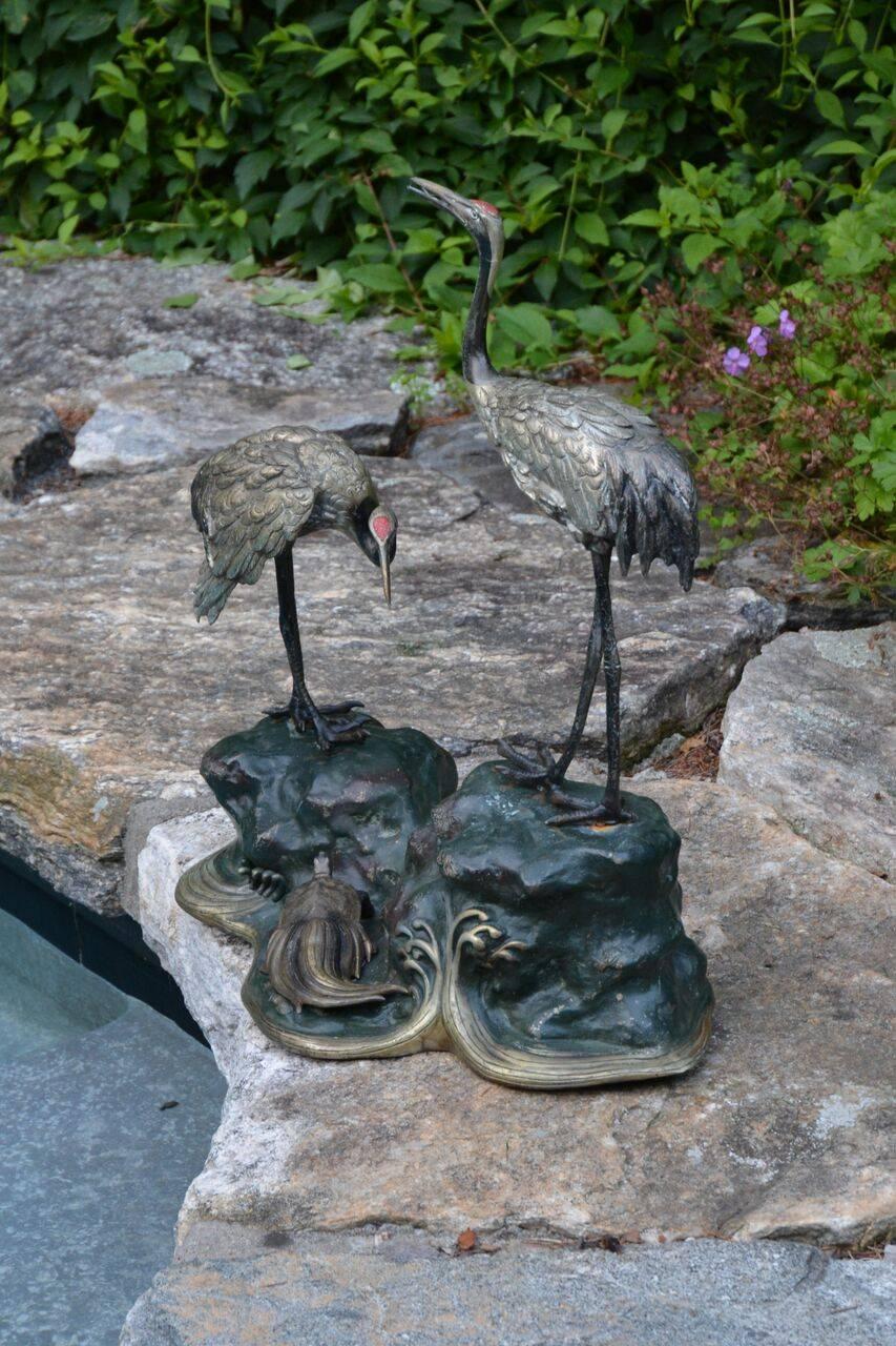 A bronze polychromed sculptural group of two red-crowned cranes, one with head lifted, the other with head down, on rockwork base, the base enveloped by stylized, swirling wave motifs, with a minogame, a Japanese mythological long-tailed turtle,