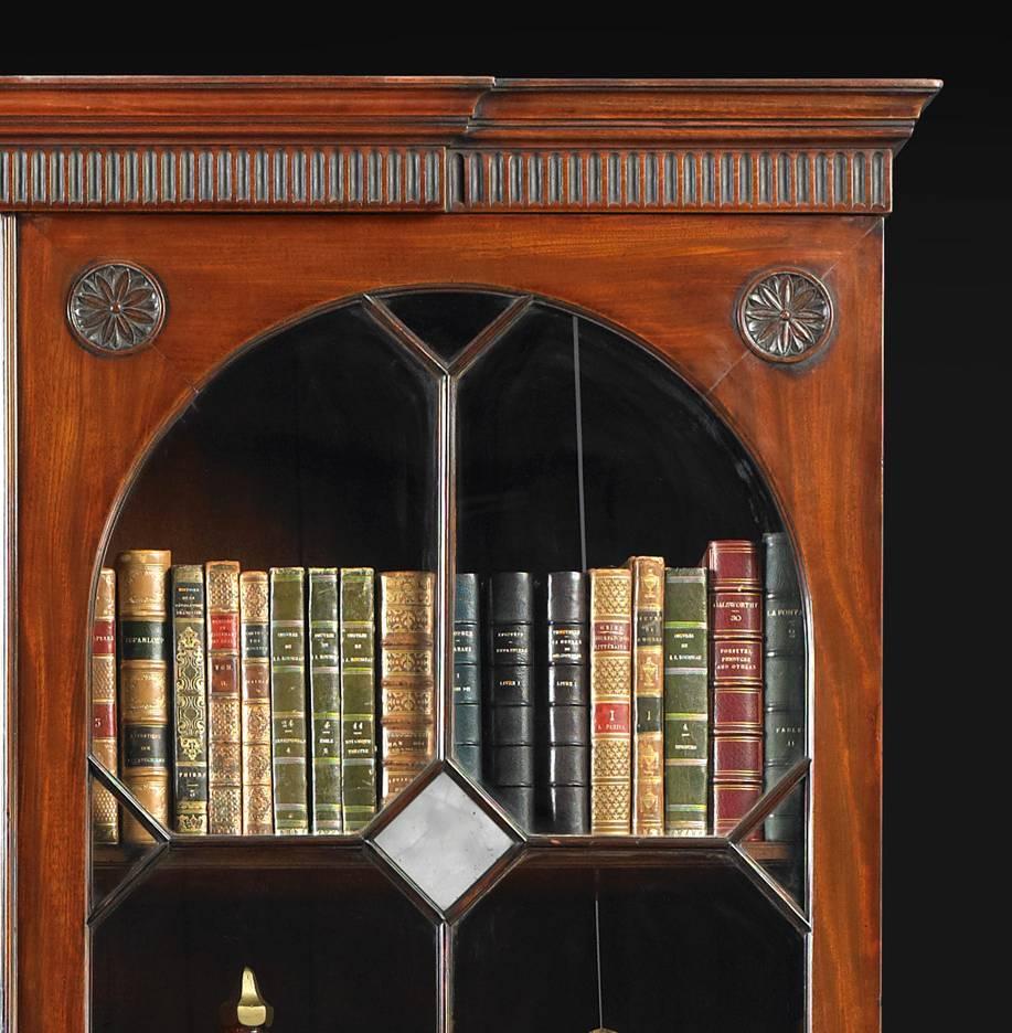 A George III mahogany bookcase, the moulded and fluted cornice above two glazed arched doors with geometric astragals and mirrored lozenges in the center, the corners with carved paterae and the lower section with four graduated drawers and all