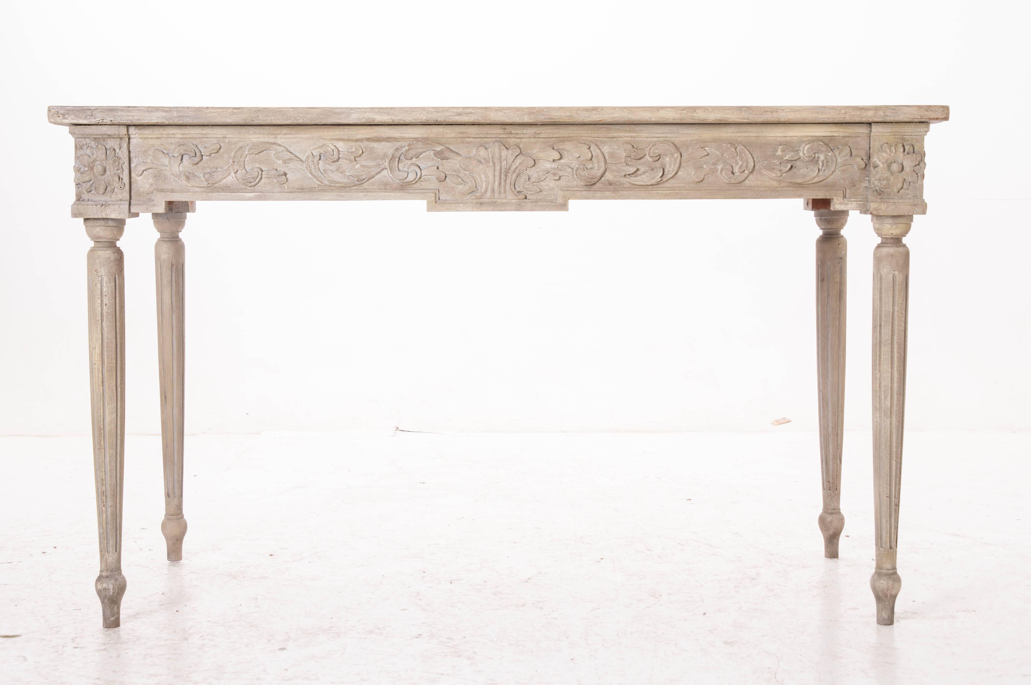 This Louis XVI style console is painted in an antique gray that bears exquisite age and patina. The apron boasts beautifully carved rosettes and scrolling acanthus leaves in deep relief. The console rests on delicate turned legs that have been