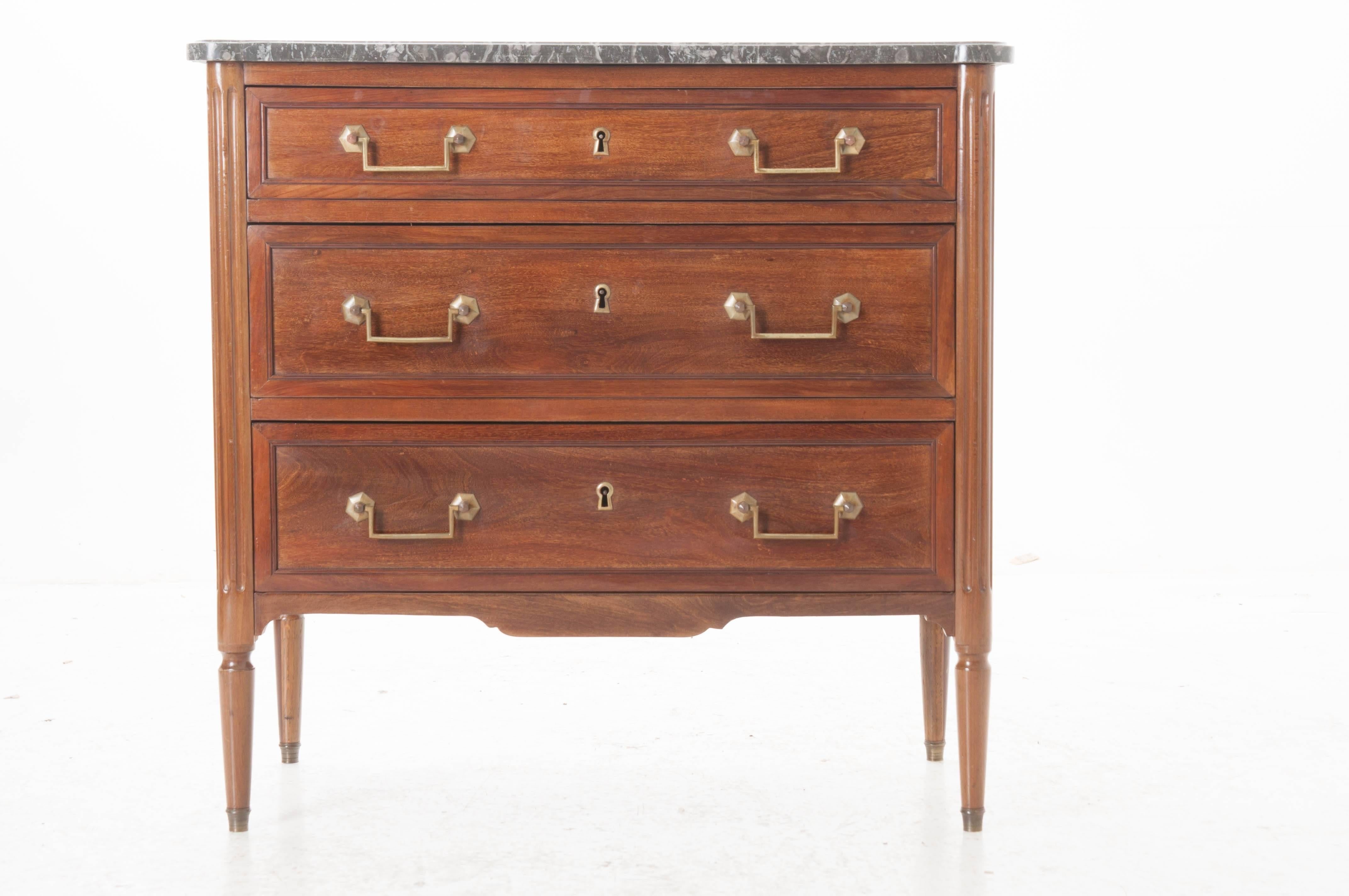 A small mahogany Louis XVI commode with original gray fossil marble top, shaped to fit the three-drawer piece. The drawers have two brass drop bail handles each and are flanked by turreted columns with fluted carving. The commode had a shaped apron
