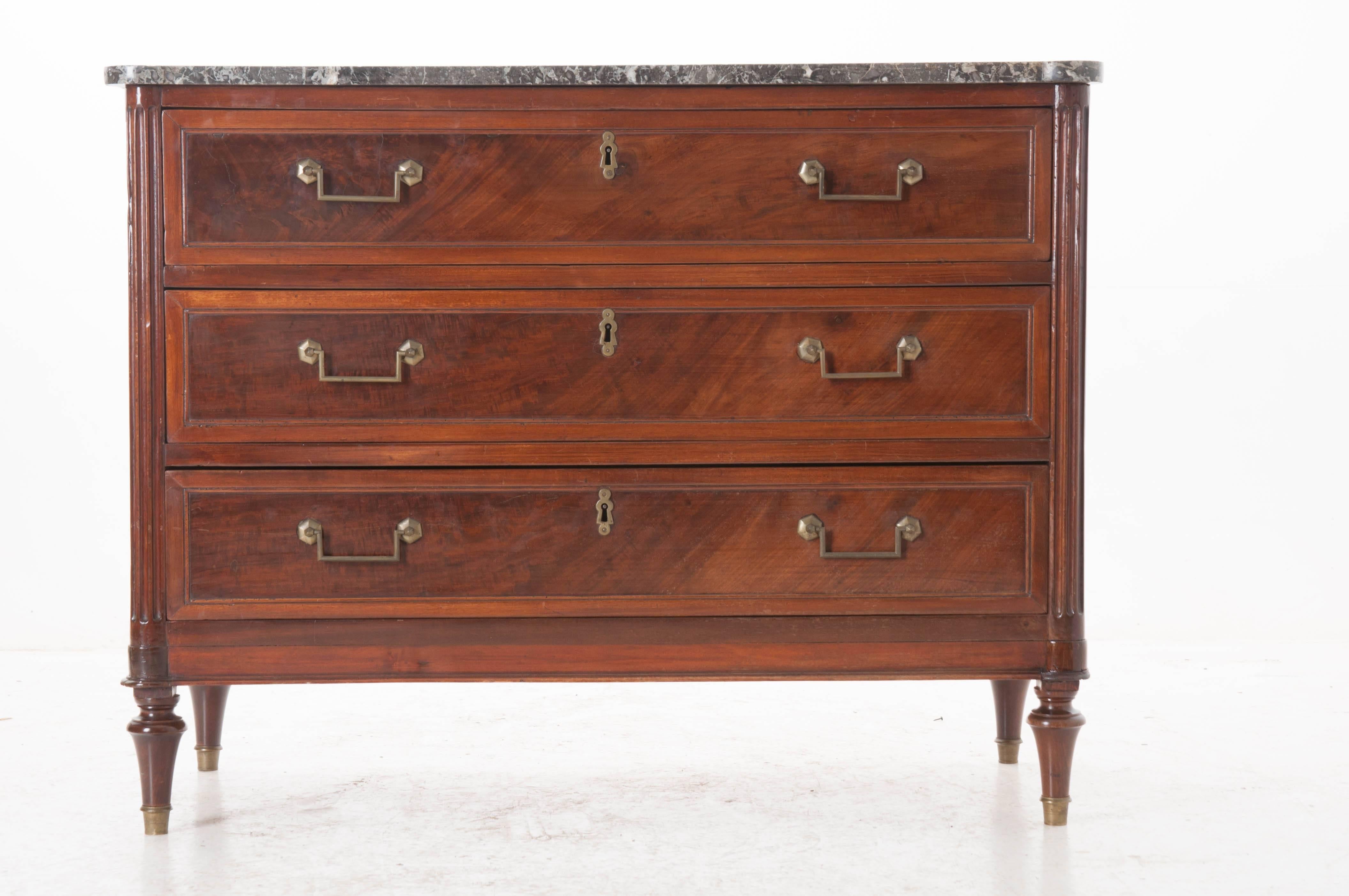 A French Louis XVI three-drawer commode with its original shaped, gray marble top. The facade is dominated by the piece's three large drawers. These are each finished with continuous, unbroken pieces of mahogany veneer and retain their original